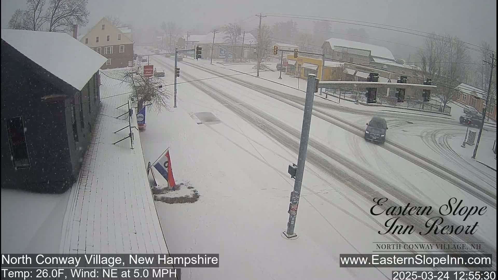 A snow-covered street scene shows a sparsely trafficked intersection with buildings on either side, under a light snowfall.