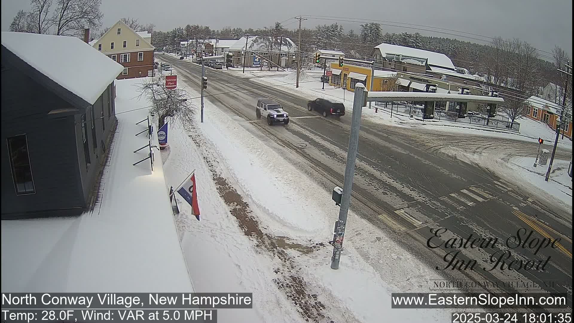 A snow-covered street scene shows two vehicles driving through a town intersection on a partly cloudy day with a temperature of 28°F and a variable wind at 5.0 mph.