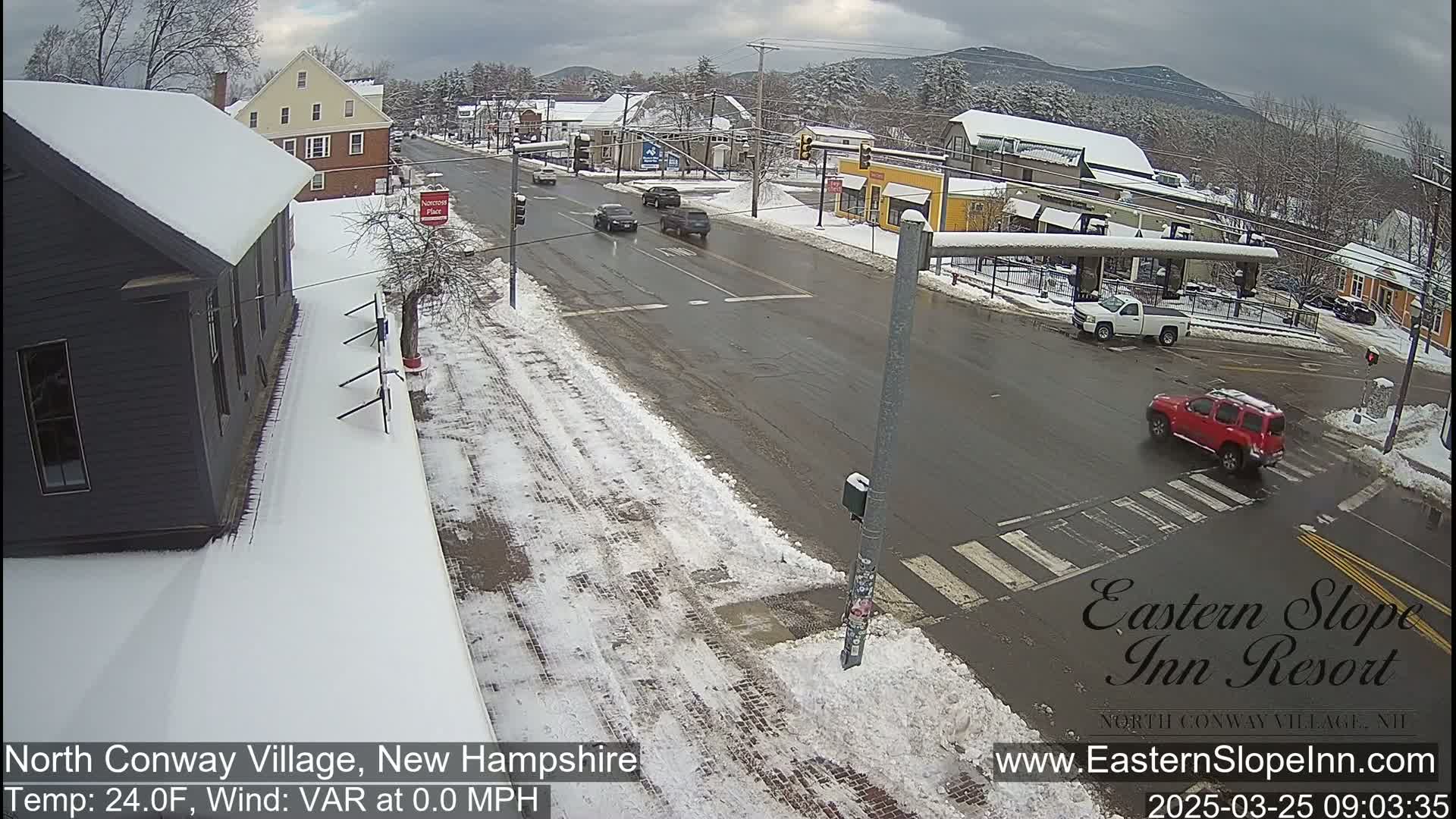 A snow-covered street scene shows several cars at an intersection in a small town, with a partly cloudy sky and mountains in the distance.