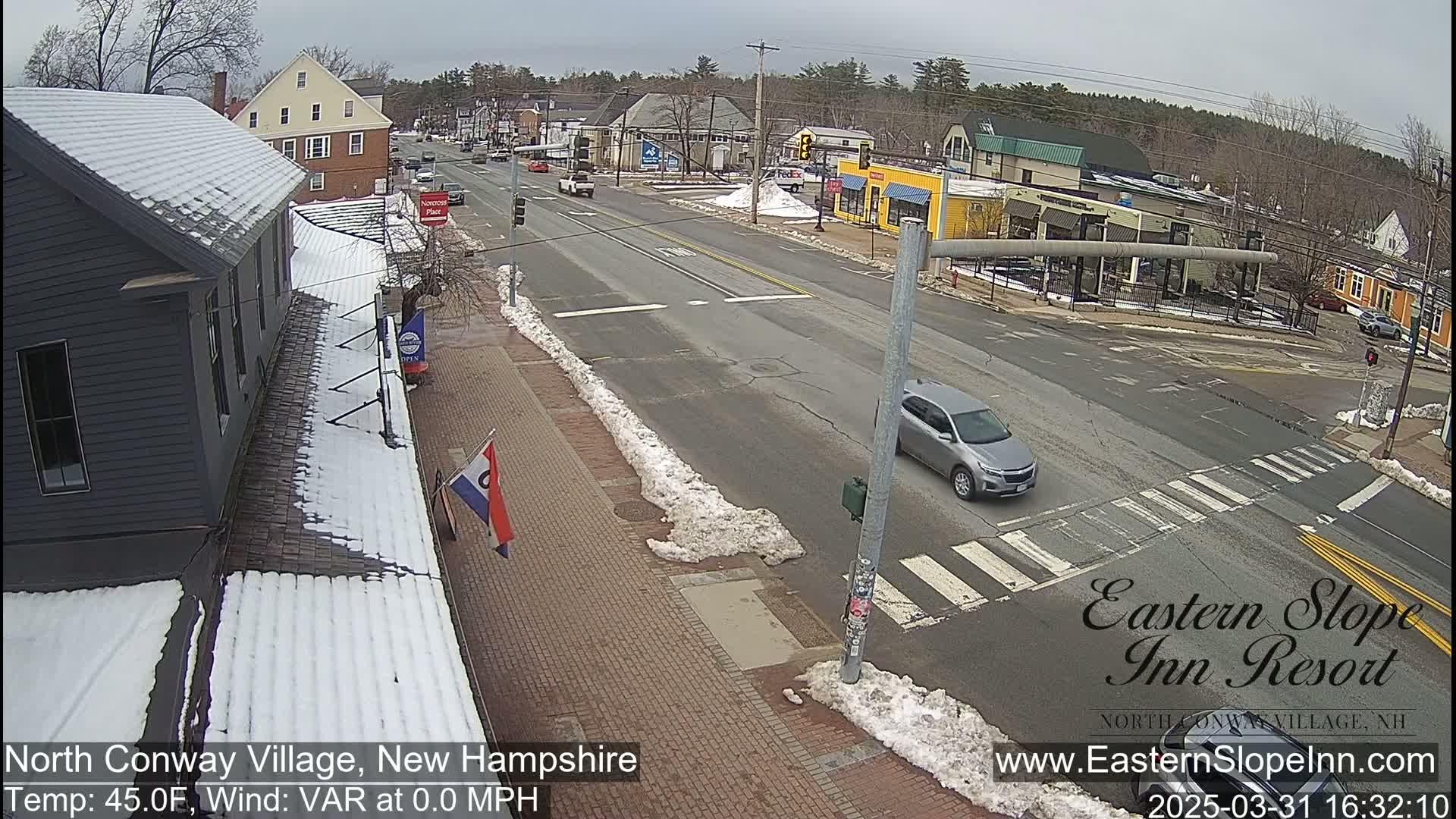 A street scene shows a partly snowy, paved road with buildings and cars under a cloudy sky.