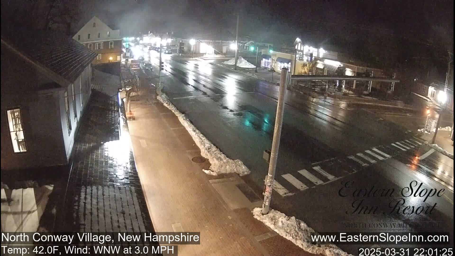 A rain-slicked street scene at night shows a town intersection with snow banks at the edges of the road and buildings lining the street under dim lighting.