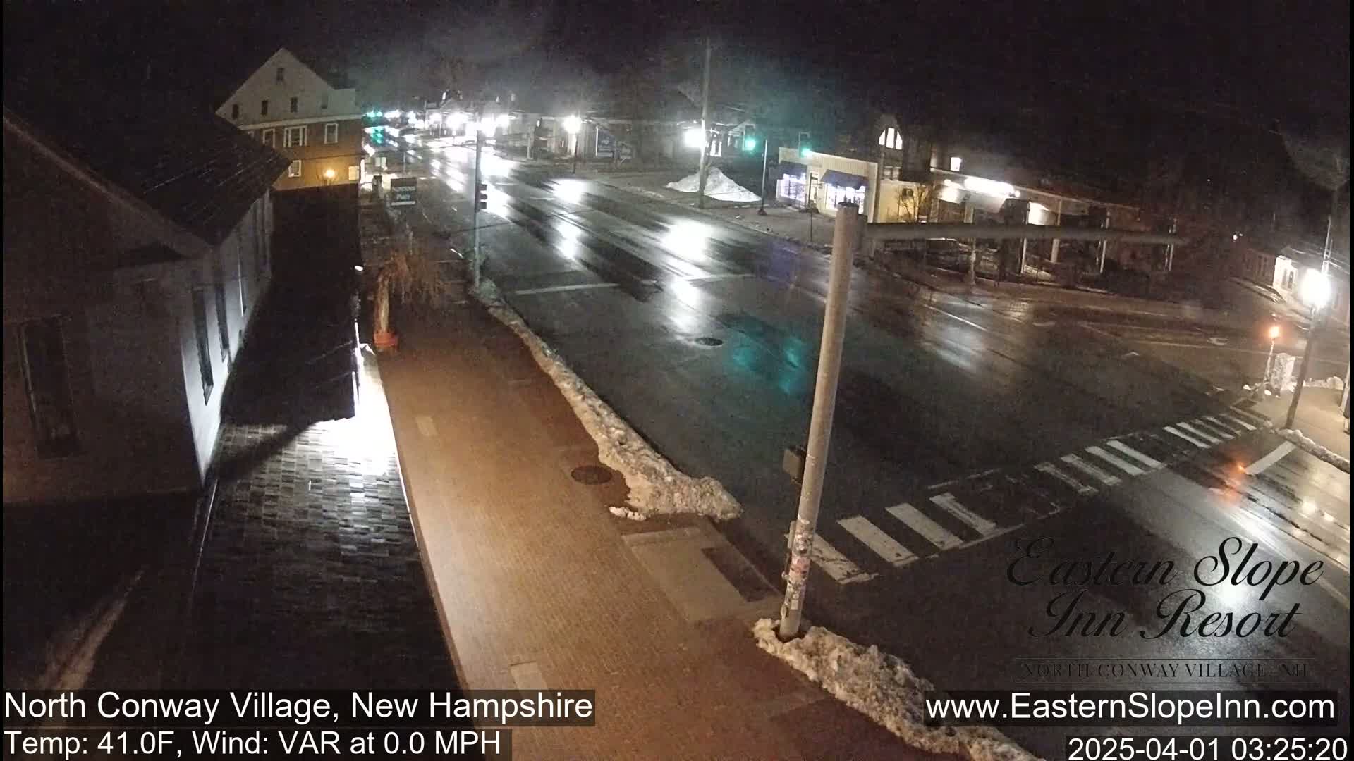 A wet, nighttime street scene shows a sparsely trafficked road with buildings and patches of snow along the curb.