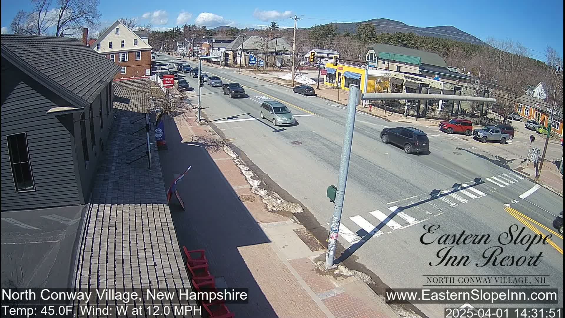 A sunny street scene with light traffic and patches of melting snow on the sidewalks, set against a backdrop of mountains under a partly cloudy sky.