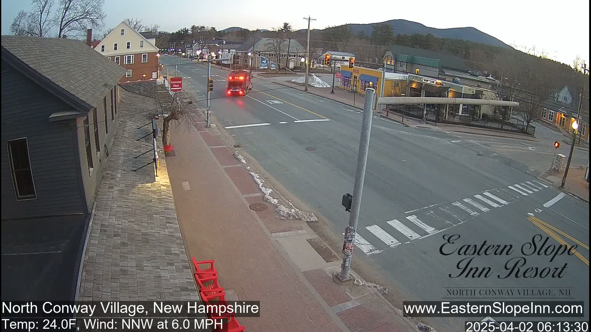 A mostly empty street intersection in a village, with a truck and some pedestrians visible, under a clear twilight sky.