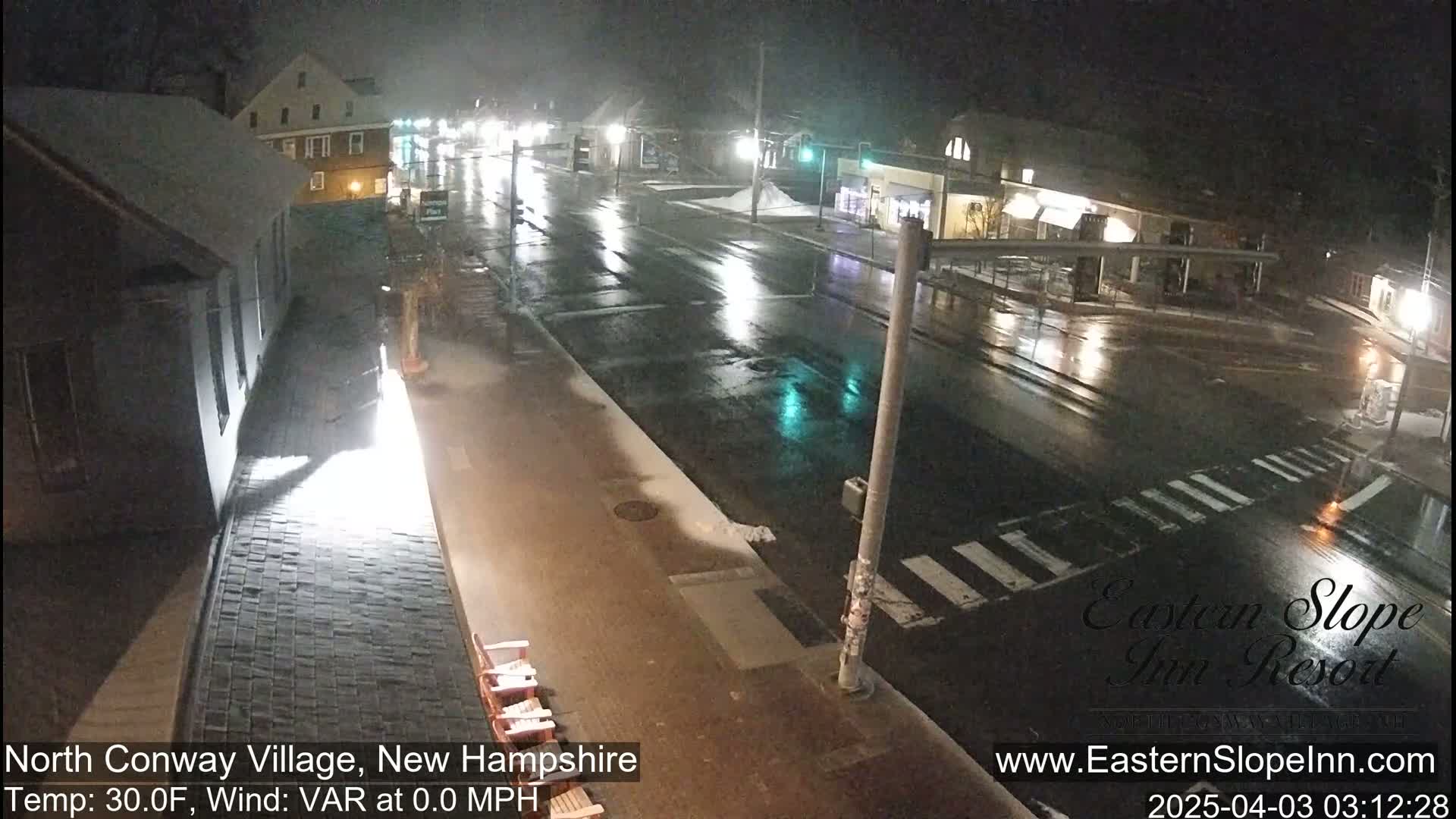 A nighttime view of a rain-slicked street in a village, with buildings lining the road and some snow patches visible on the sidewalk.