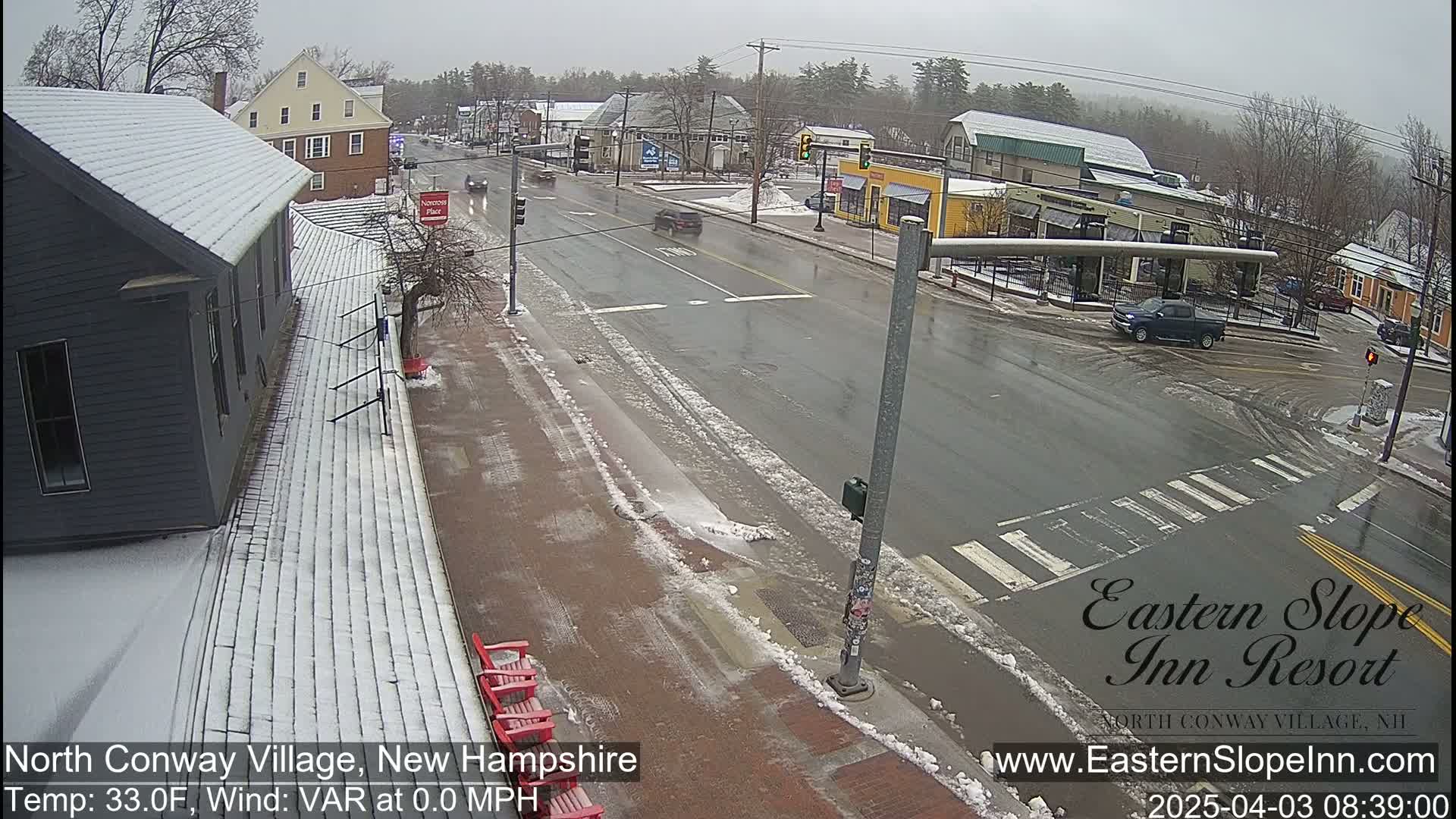 A snow-covered street scene shows a partly sunny day with a few cars driving, some buildings, and red Adirondack chairs.
