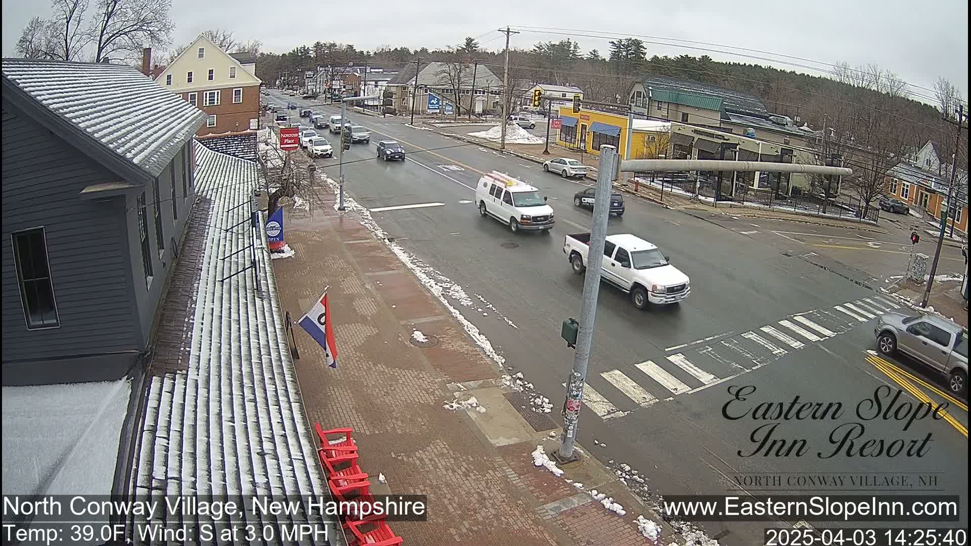 A street scene on an overcast day shows several vehicles at an intersection, with patches of snow and ice visible on the ground and rooftops.