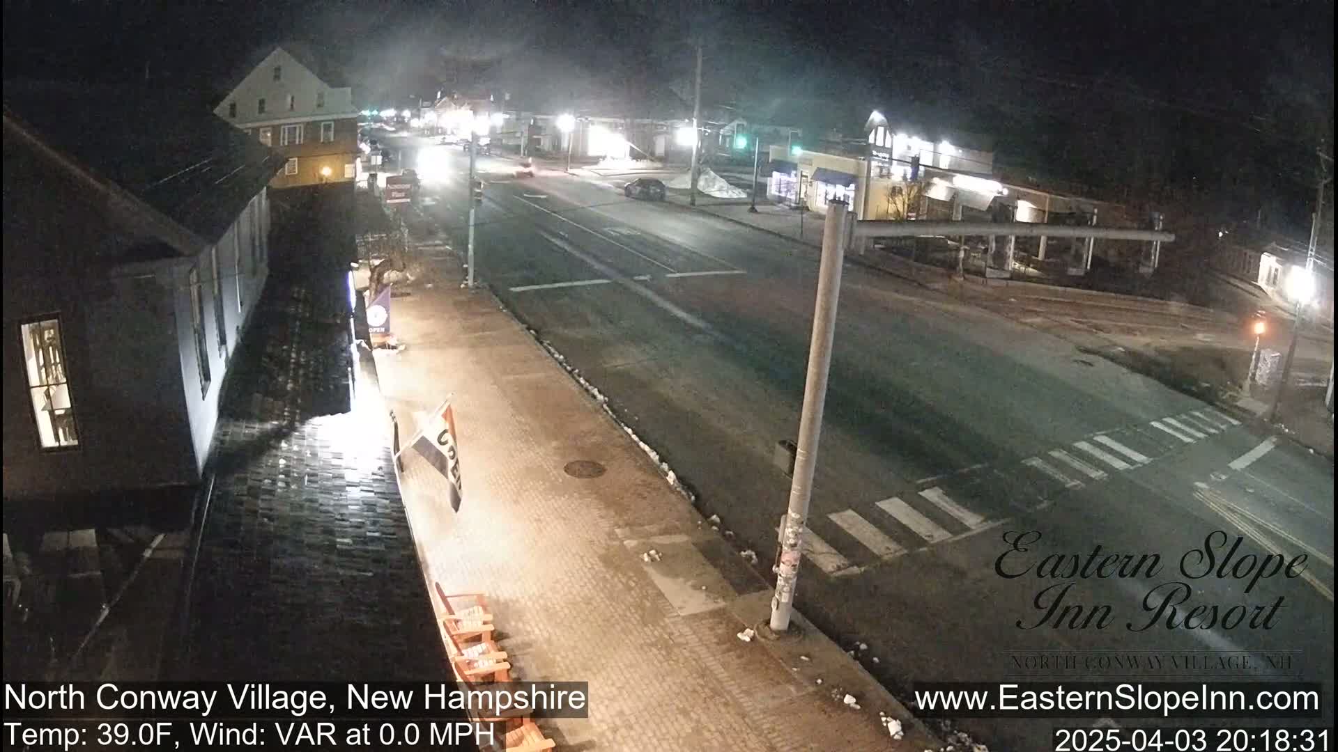 A nighttime street scene shows a mostly empty road and sidewalks with some snow patches, viewed from an elevated position.