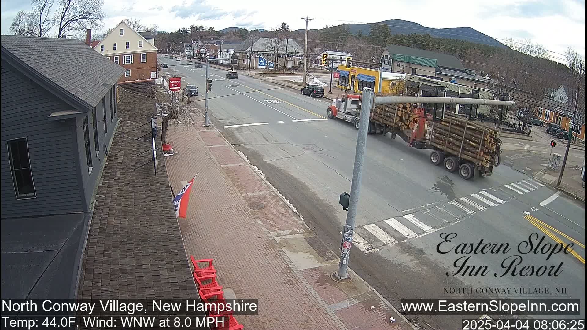 A semi-truck carrying logs drives through a town intersection on a partly cloudy day with an 8 mph wind.
