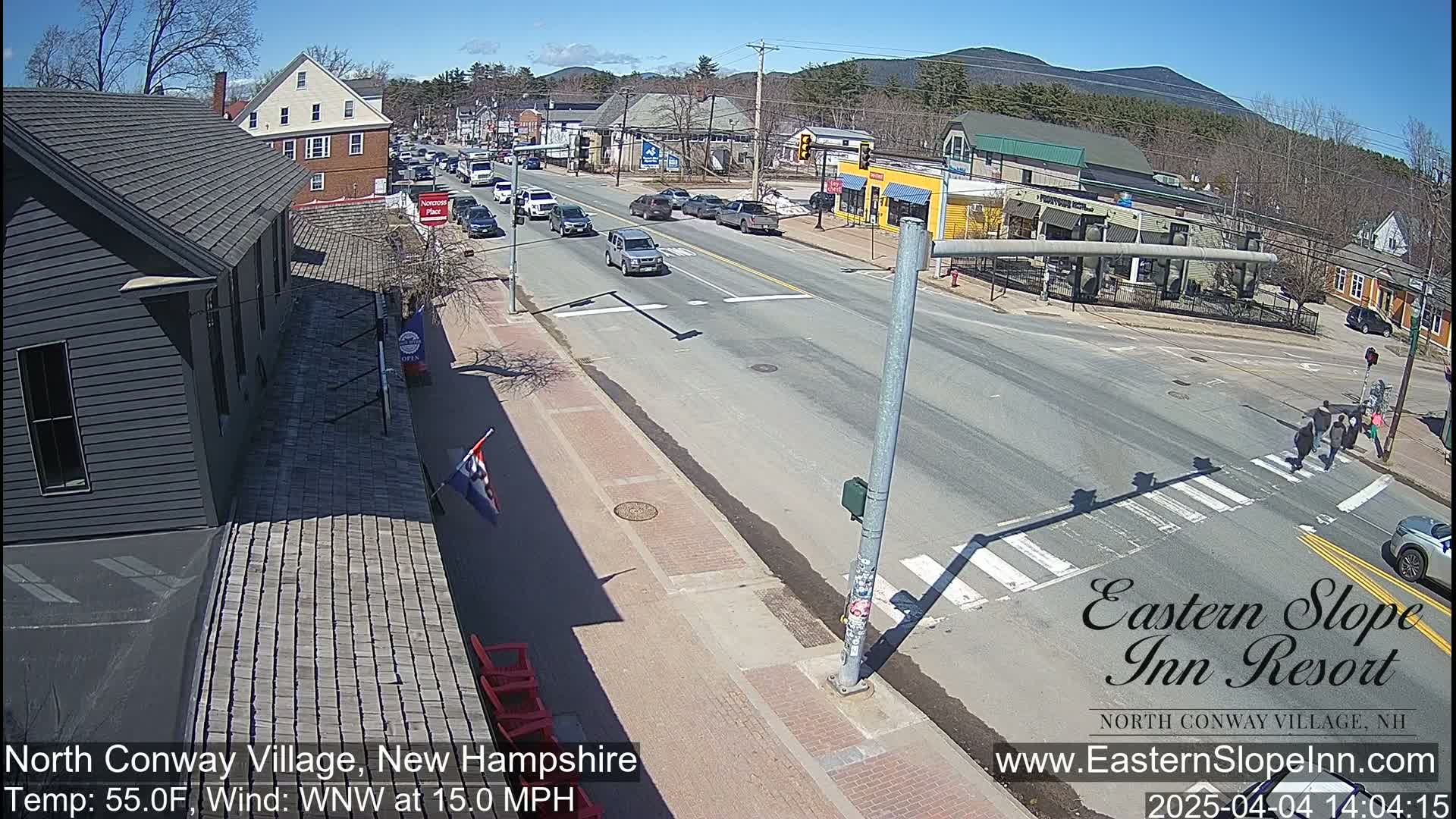 A sunny, partly cloudy day shows a street scene with several cars and pedestrians crossing at an intersection, with buildings and mountains visible in the background.