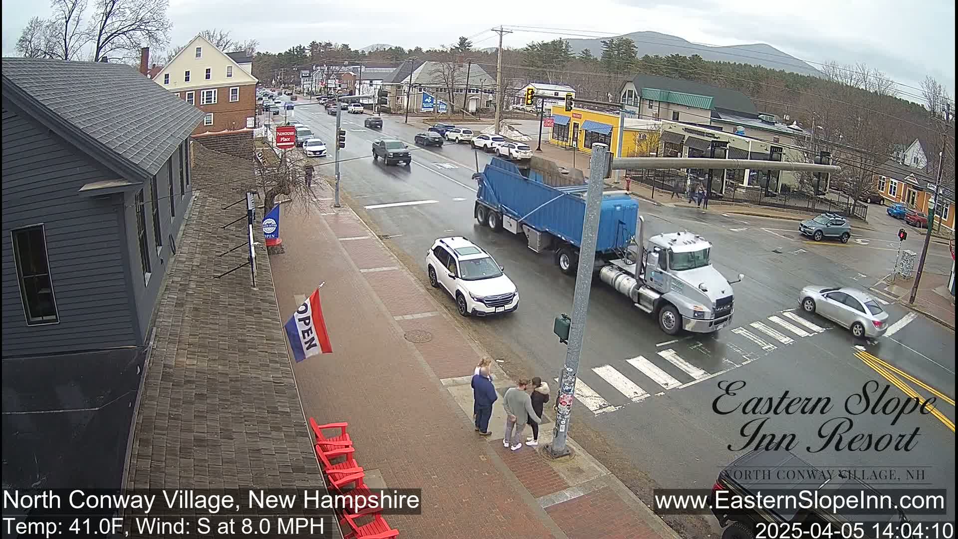 A wet street scene in a village shows several cars and trucks, pedestrians, and a group of people standing on a sidewalk under a cloudy sky.