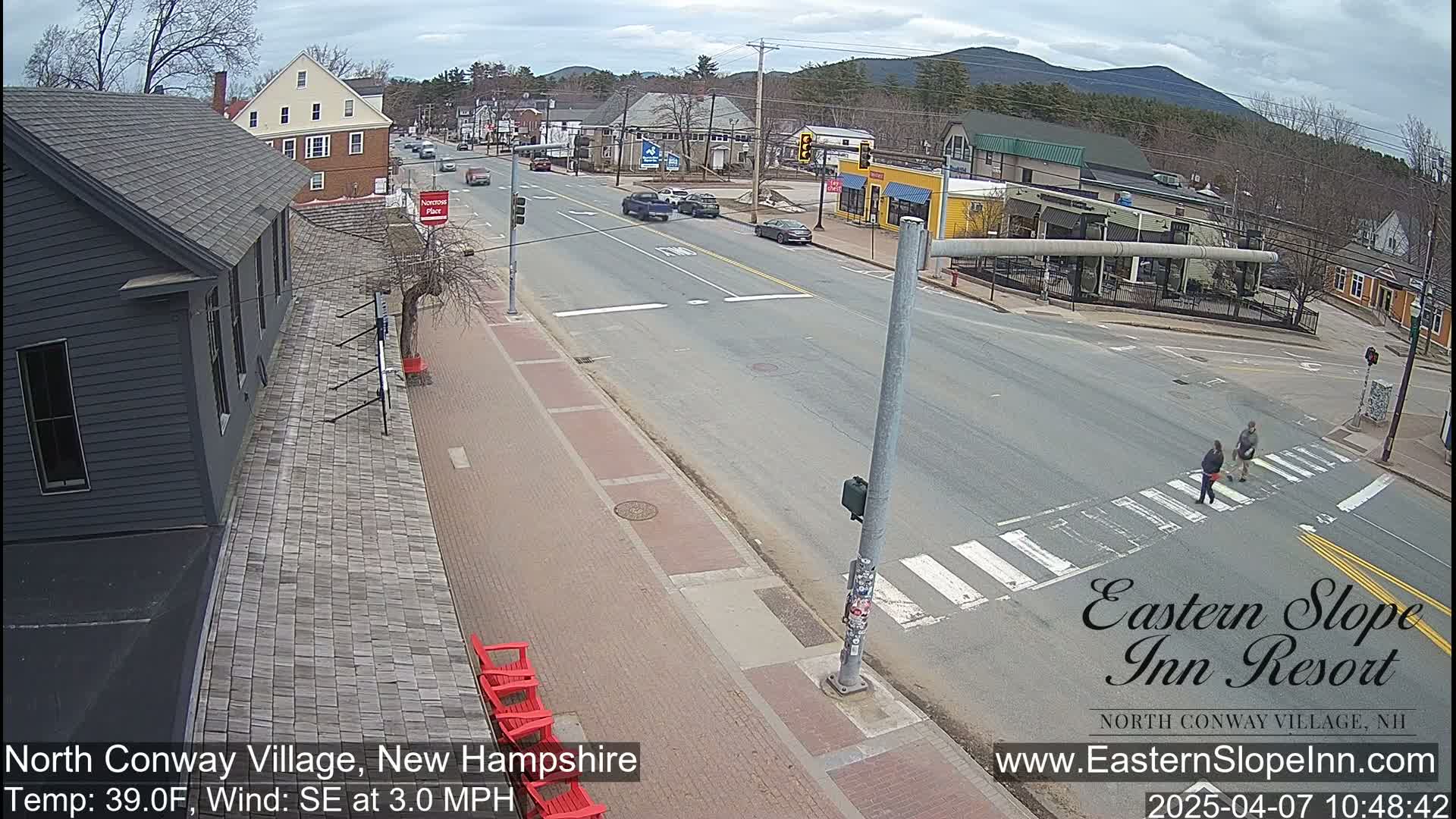 A street scene on a partly cloudy day shows a road with cars and pedestrians, brick sidewalks with red chairs, and buildings including a dark-colored building on the left, and other buildings and businesses along the road.