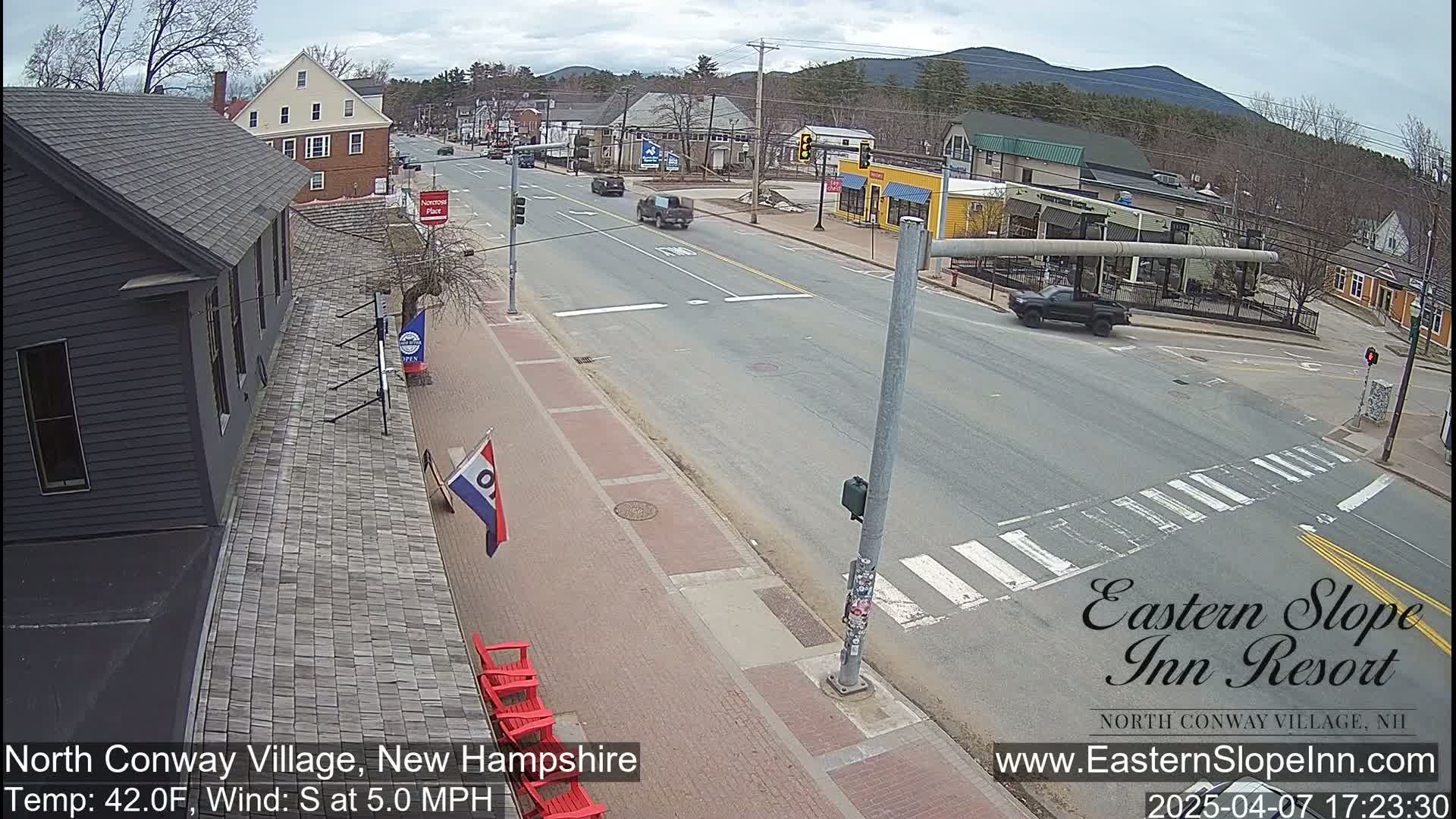 A mostly sunny street scene shows a town intersection with several buildings, some cars, and red Adirondack chairs, under a partly cloudy sky with a south wind.