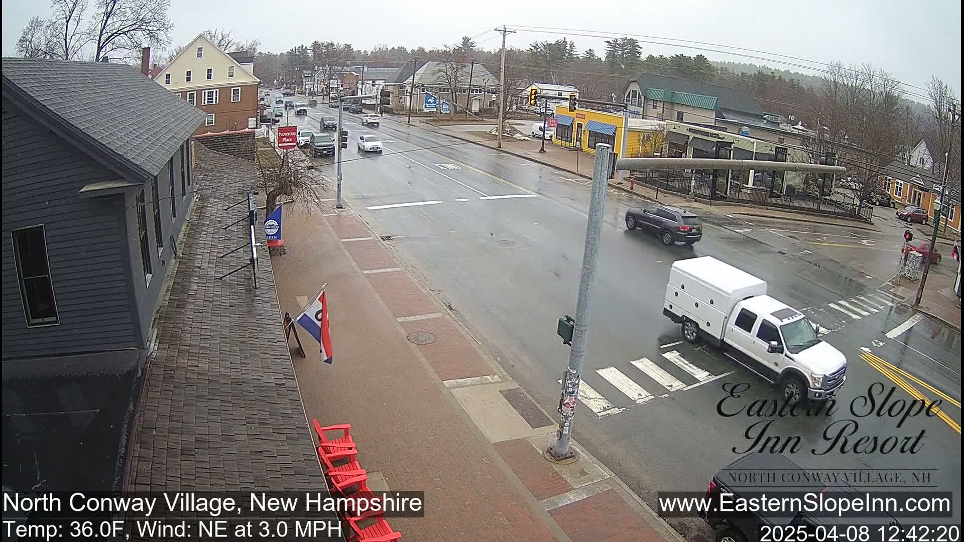 A wet street scene shows a village intersection with several cars and a white truck, under a gray, overcast sky.
