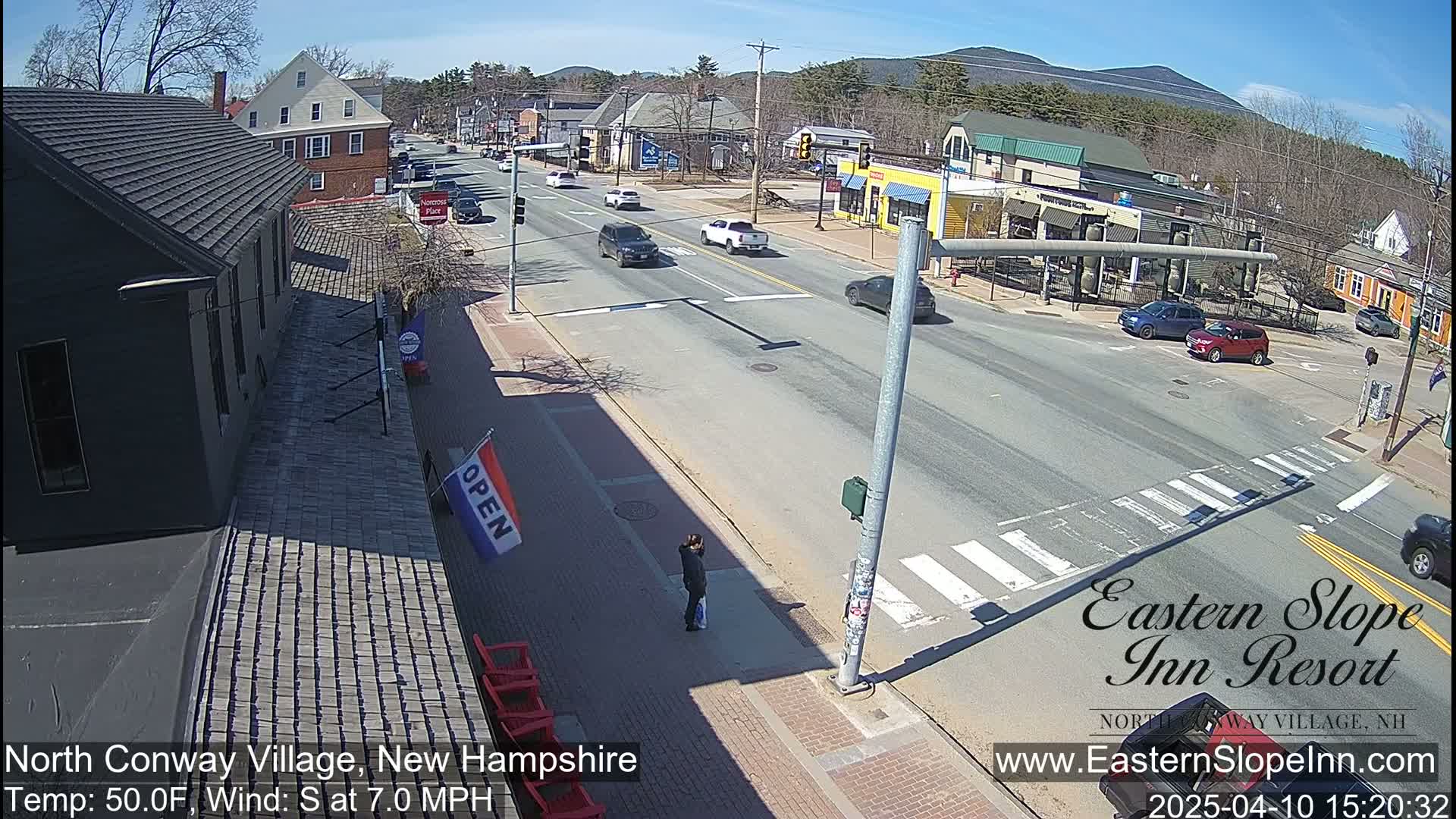 A sunny day reveals a street scene in a village, with several cars on the road and pedestrians on the sidewalk, alongside buildings of varying styles and sizes, and mountains in the background.
