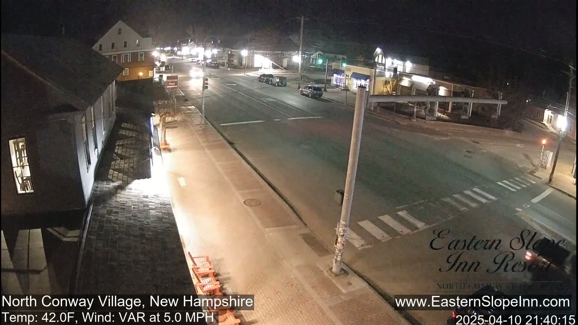 A nighttime street scene shows a mostly empty road with a few cars, buildings, and a crosswalk under a clear sky.