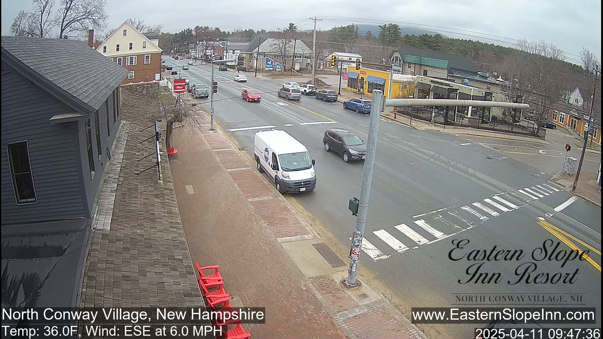 A street scene on an overcast day shows a mostly empty road with several cars and a white van, alongside brick sidewalks and buildings, including a dark gray building with red chairs outside.