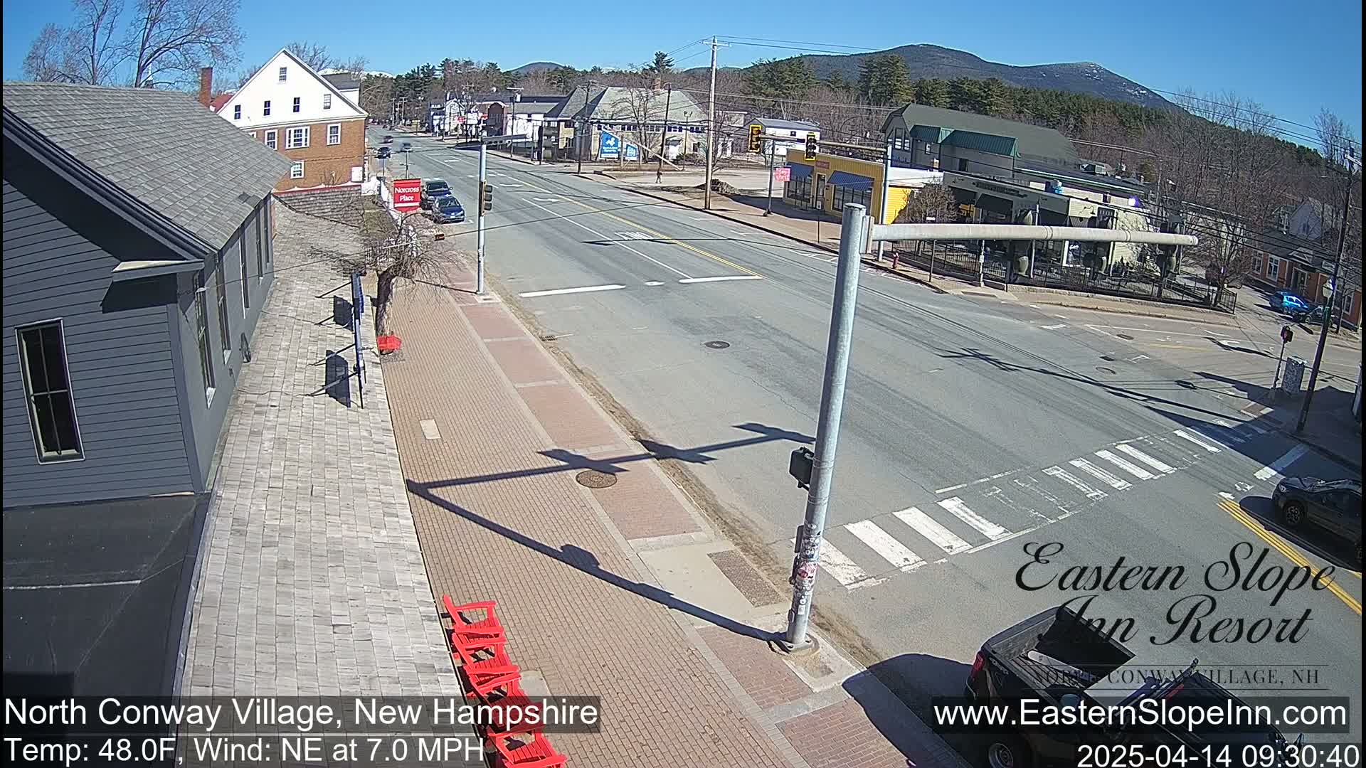 A sunny, partly cloudy day shows a view of a town's main street intersection with cars and buildings, and red chairs on a brick sidewalk.