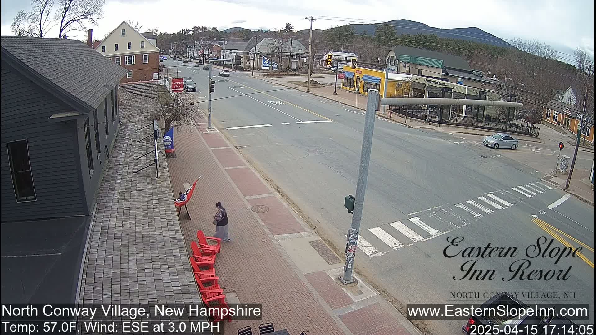 A mostly sunny street scene in a small town shows a car at an intersection, a pedestrian walking on a sidewalk with red chairs, and a person relaxing in a hammock outside a building.