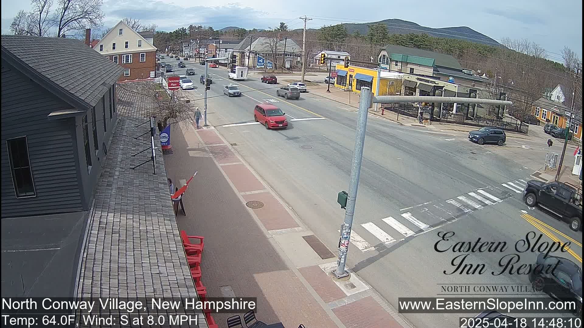 A street scene on a partly sunny day with a light south wind shows several cars driving through a town intersection, with buildings and pedestrians visible along the sidewalks.