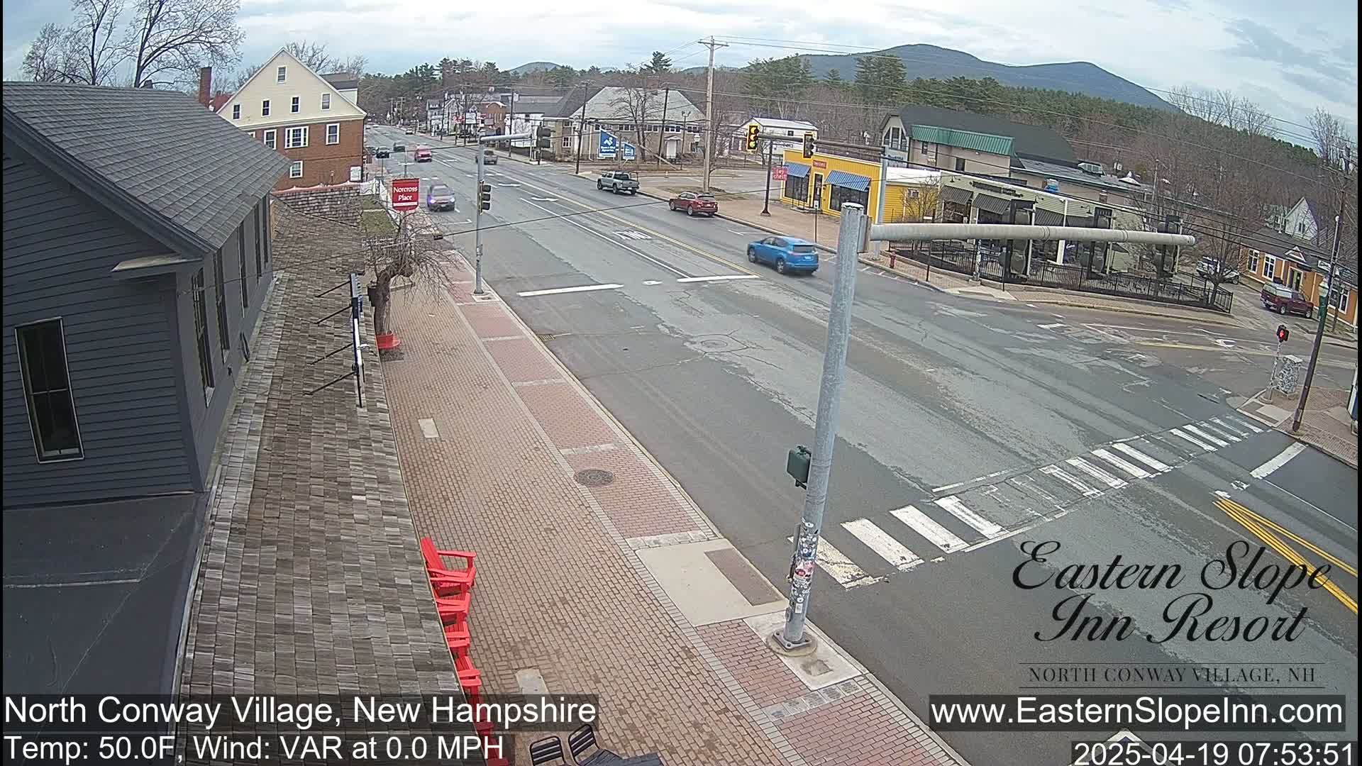 A mostly sunny view of a street intersection in a small town, with several cars and buildings visible.