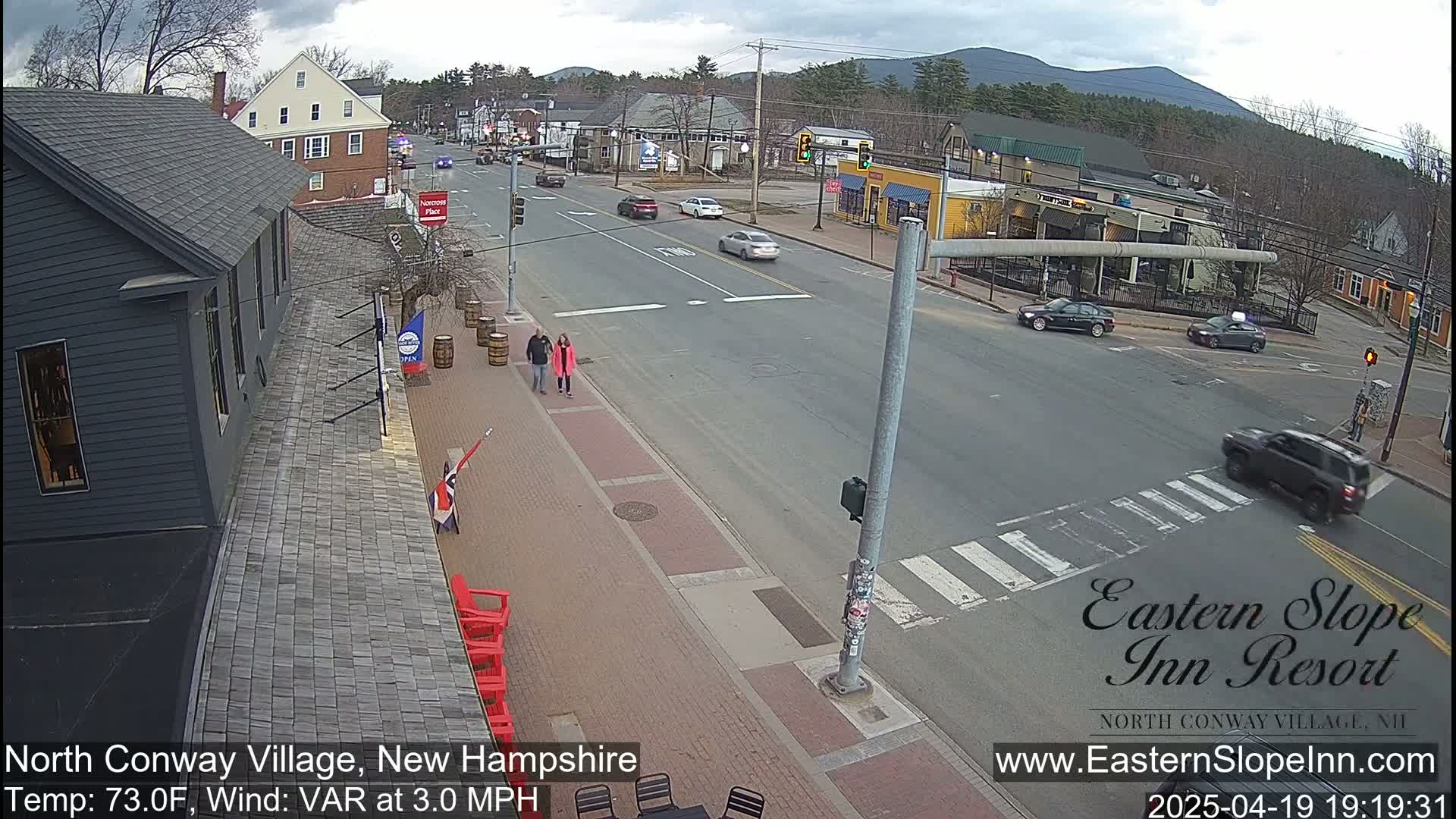 A street scene on a partly cloudy day shows a village street with cars and pedestrians, and mountains in the distance.