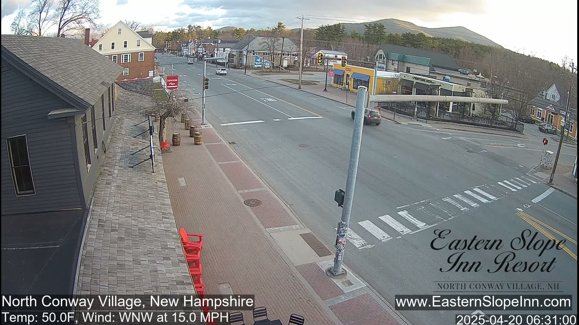 A mostly cloudy day shows a mostly empty street in a town with some buildings and trees, and mountains in the distance.