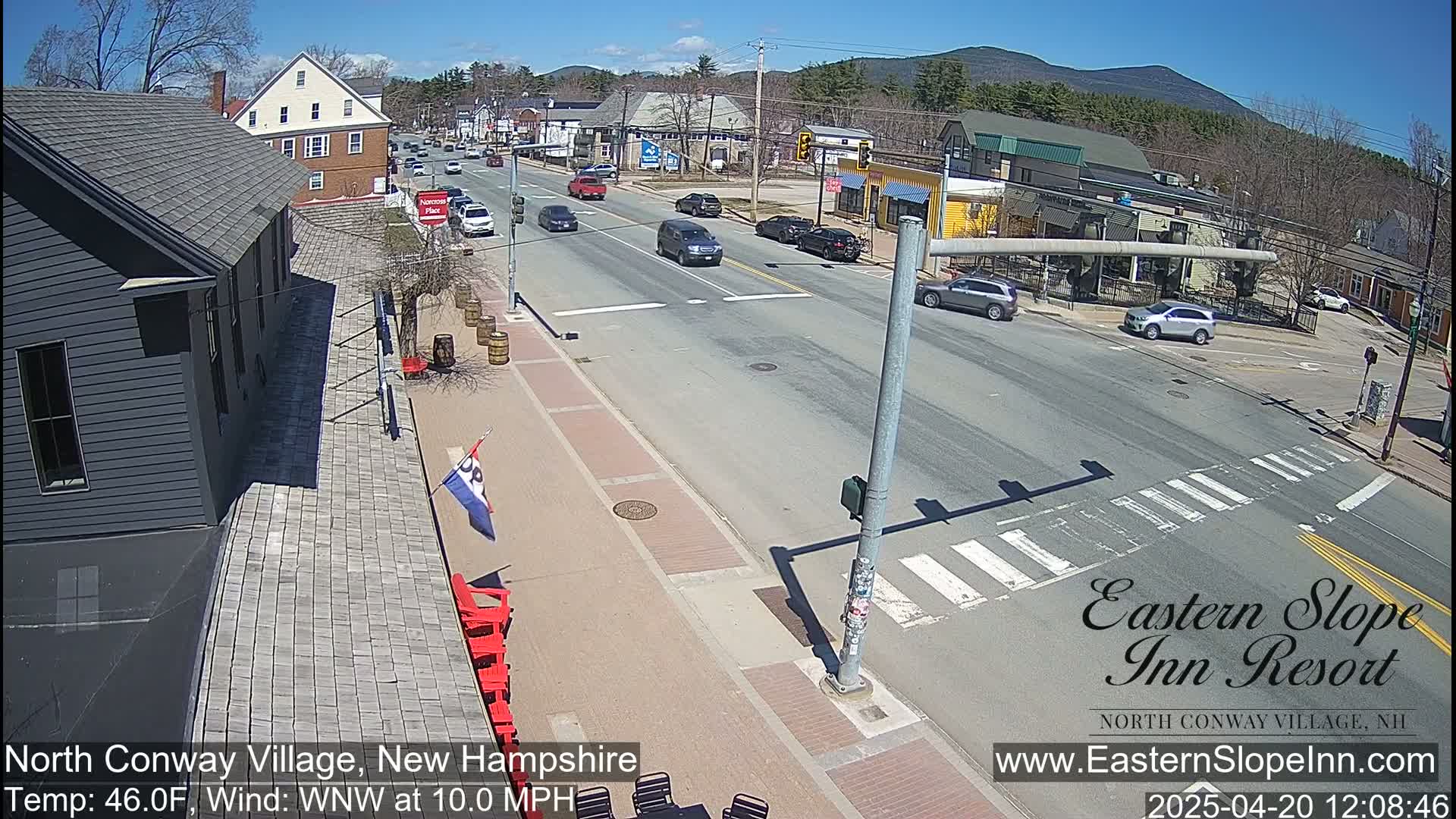 A sunny day reveals a street scene in a village with several cars on a road, buildings lining the street, and a few pedestrians visible in the distance.