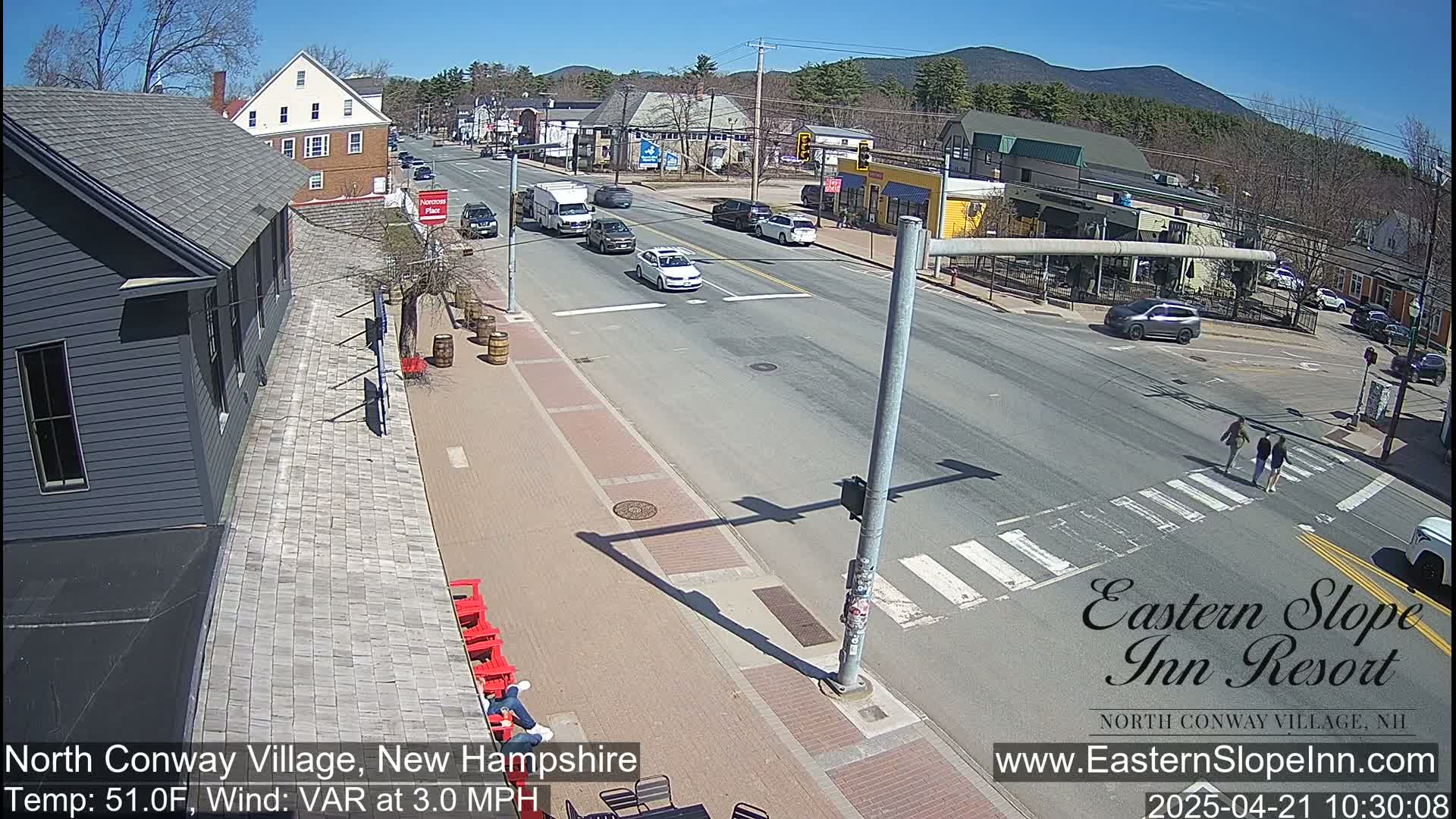 A sunny day shows a street scene with several vehicles, pedestrians crossing a crosswalk, and buildings lining the road, mountains in the background.