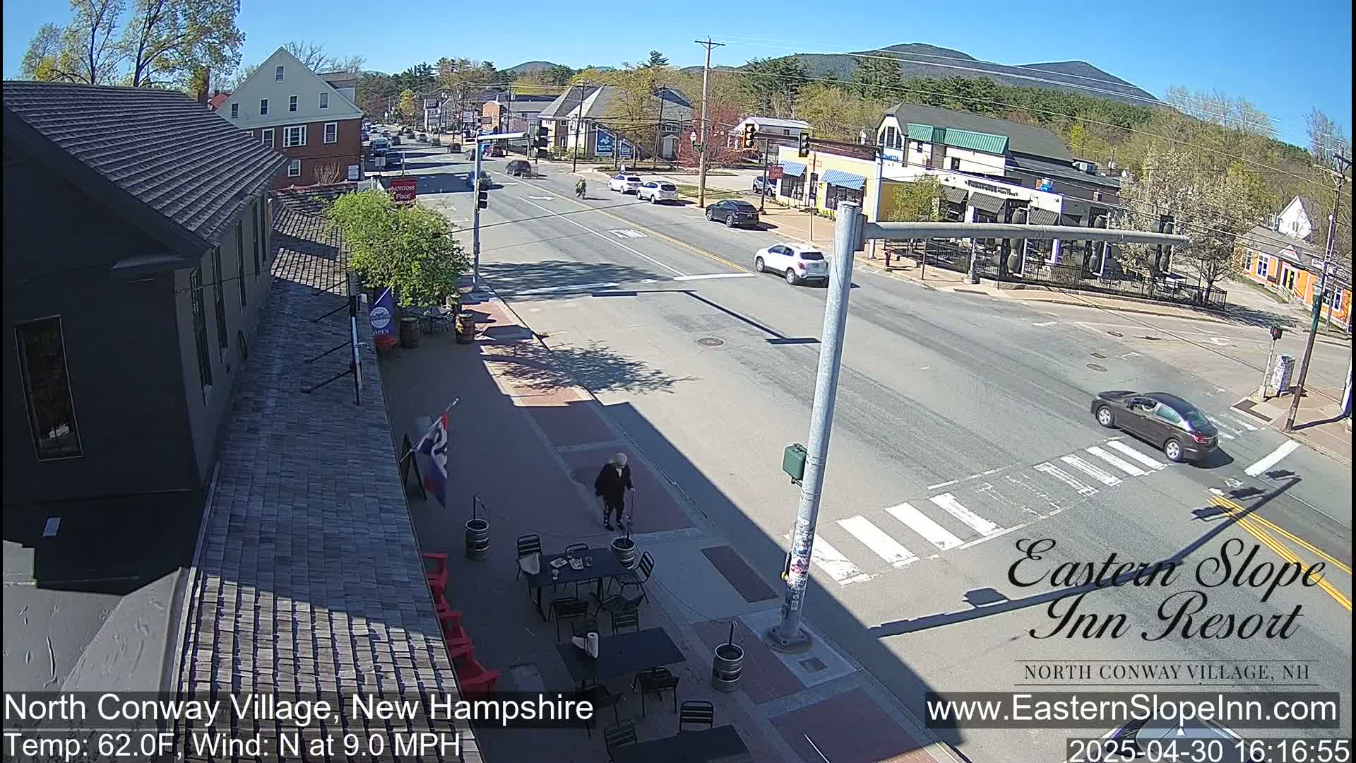 A sunny street scene in a village shows cars driving past buildings and a pedestrian walking near outdoor seating.