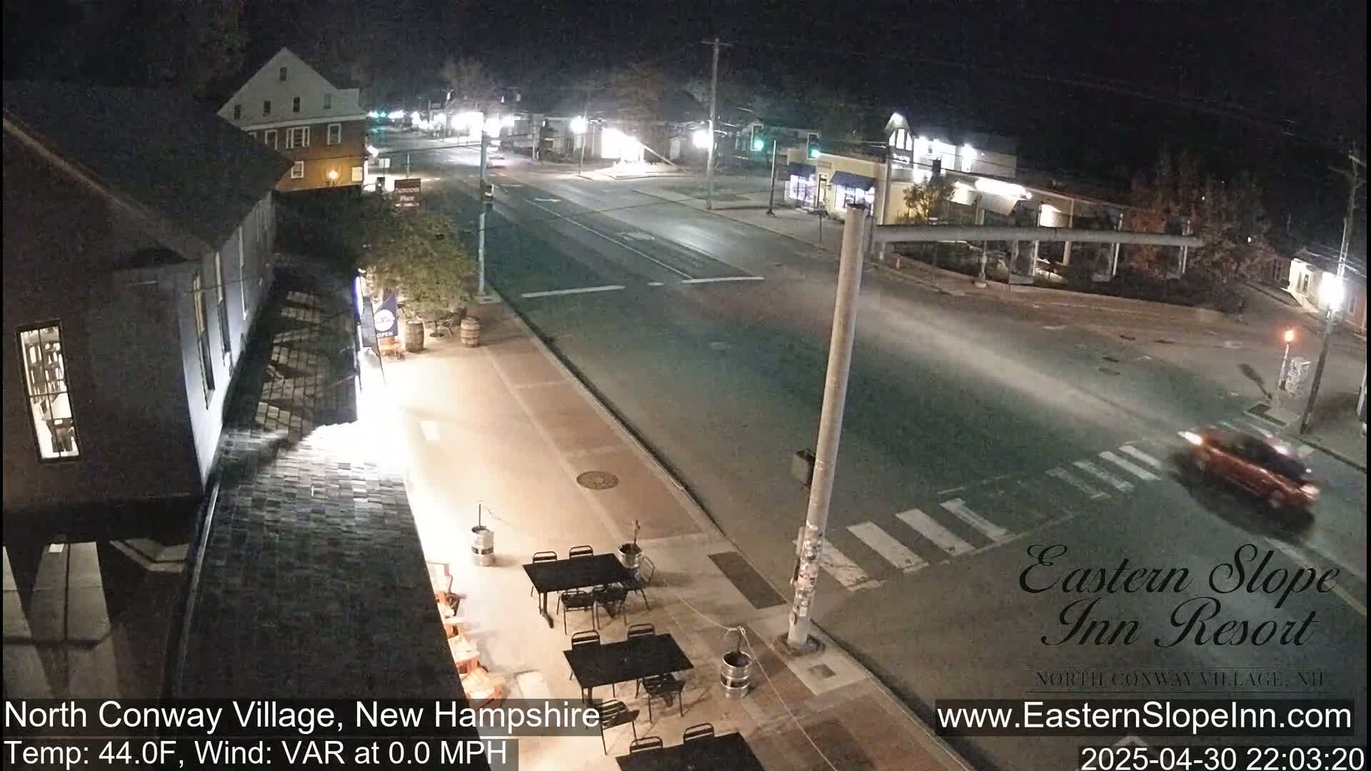 A nighttime street scene shows a mostly empty road intersecting another road, with a few buildings and an outdoor seating area visible under a clear sky.