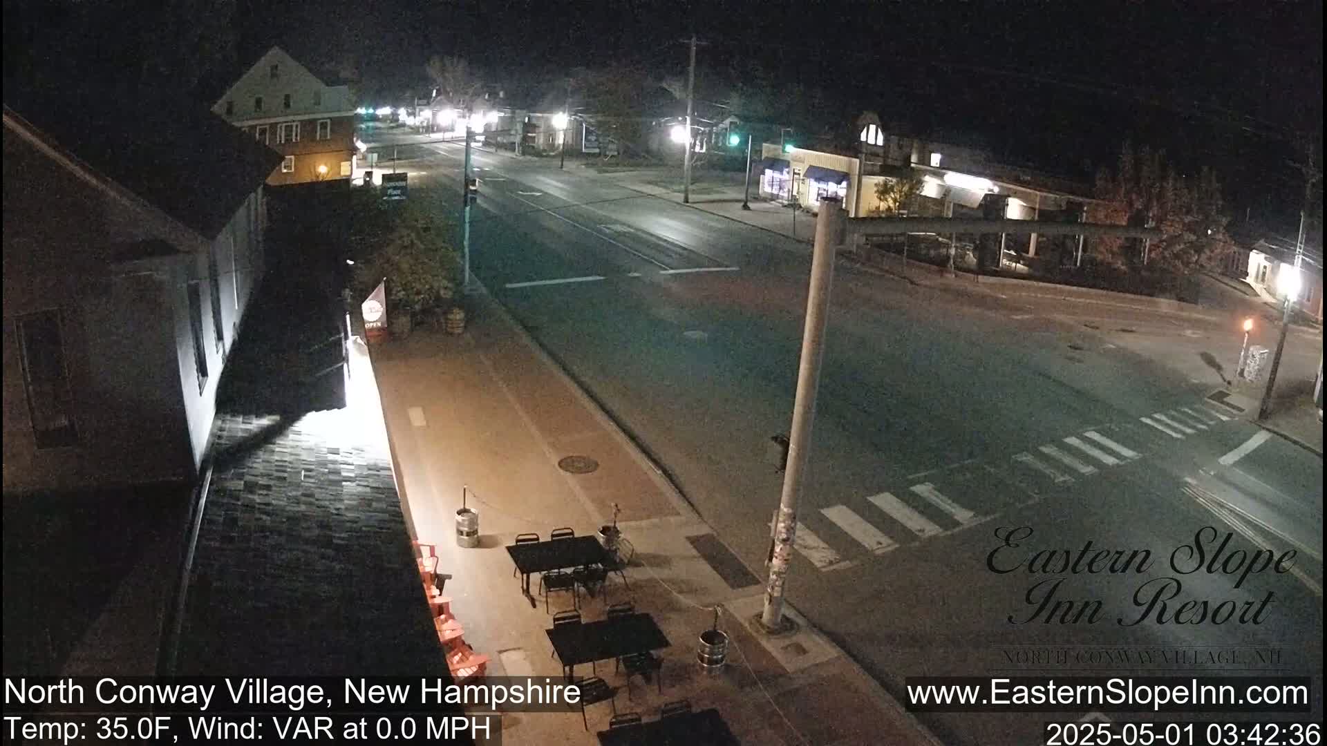 A nighttime street scene shows a mostly empty road with a few buildings visible, and some outdoor tables and chairs in front of one building.