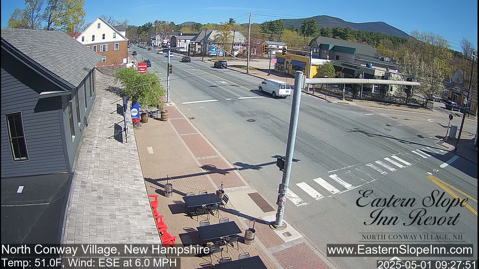 A sunny street scene shows a mostly empty road with a few cars and a white van at an intersection in a town with several buildings and outdoor patio furniture.