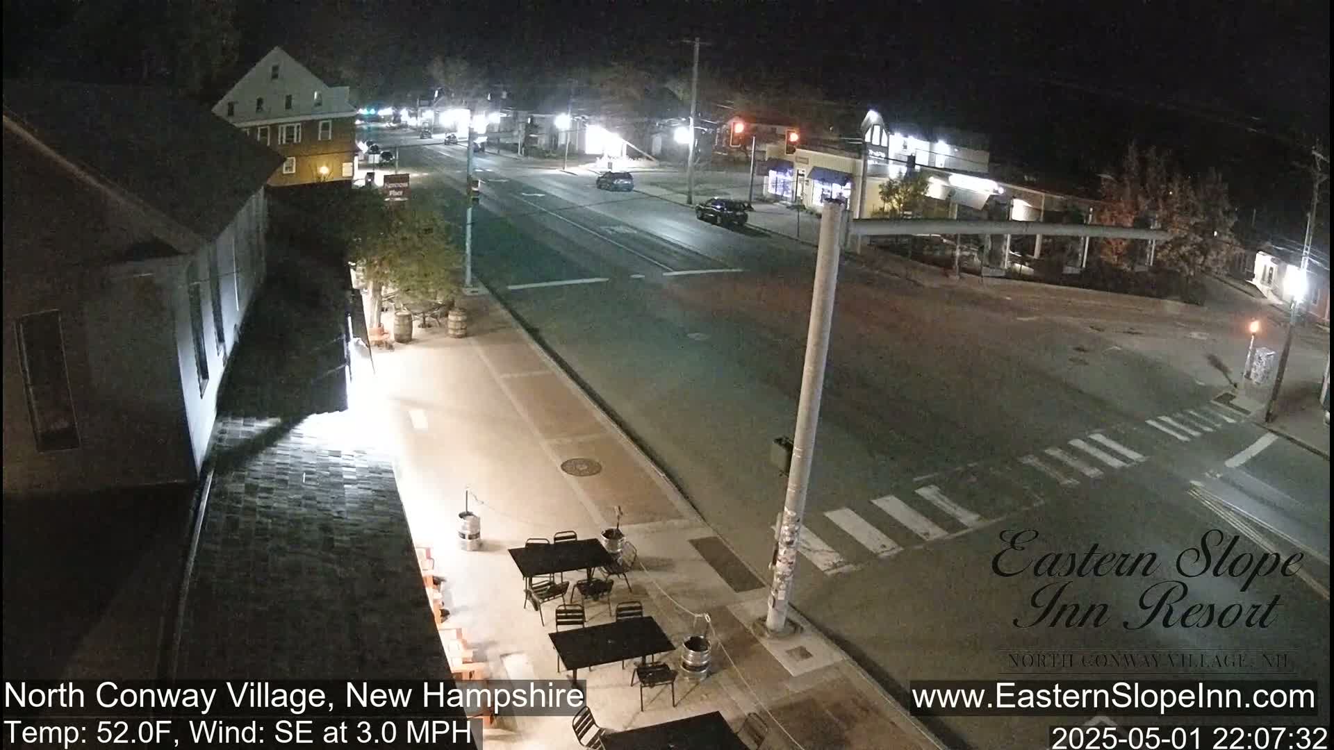 A nighttime street scene shows a mostly empty road and crosswalk, with a few cars, and an outdoor patio area with tables and chairs.