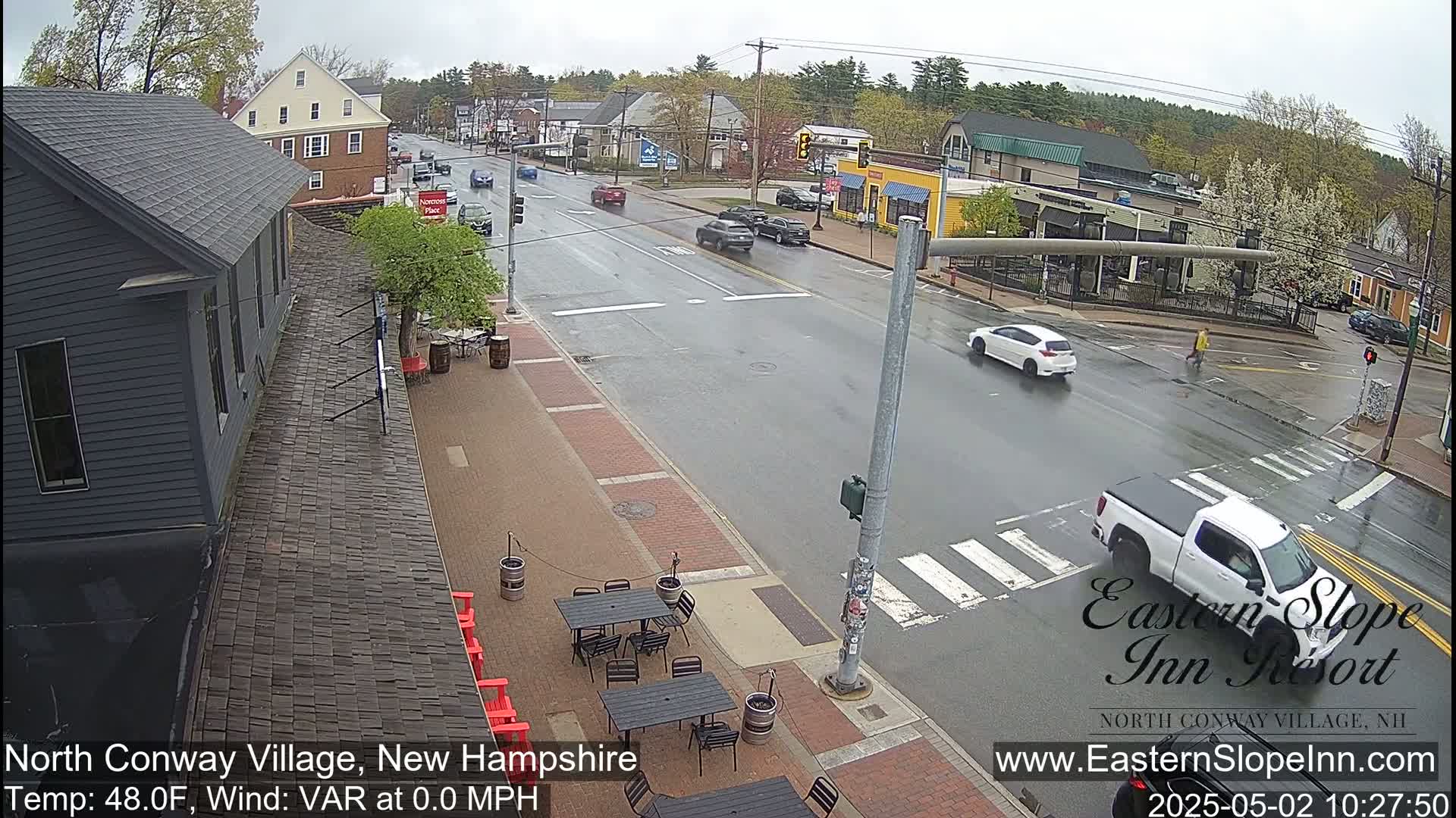 A rain-slicked street scene shows several cars driving past buildings and a sidewalk cafe with empty tables under a cloudy sky.