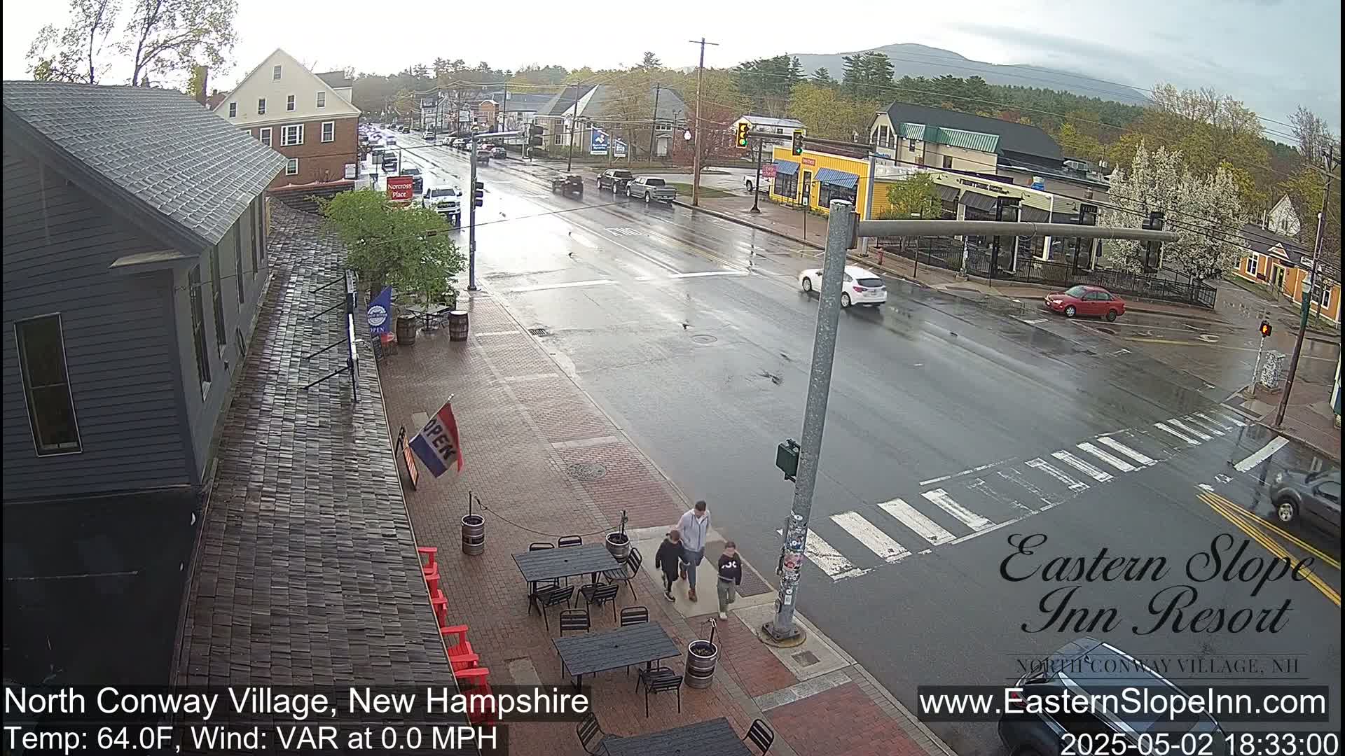 A rain-slicked street scene shows a town intersection with pedestrians crossing and cars driving, with buildings and outdoor seating visible.