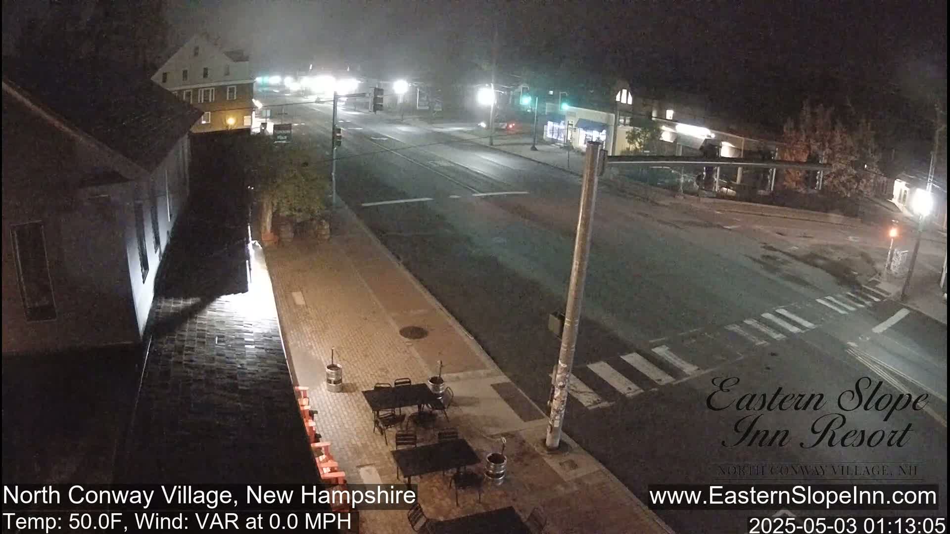 A nighttime, foggy view of a mostly empty street in a town, showing a building and outdoor seating area to the left and other buildings across the road.