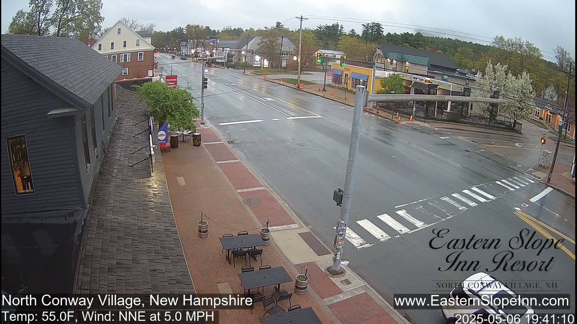 A wet street scene shows a brick-paved sidewalk with outdoor seating, a dark-colored building, and a crosswalk in front of several other buildings, all under a cloudy sky.