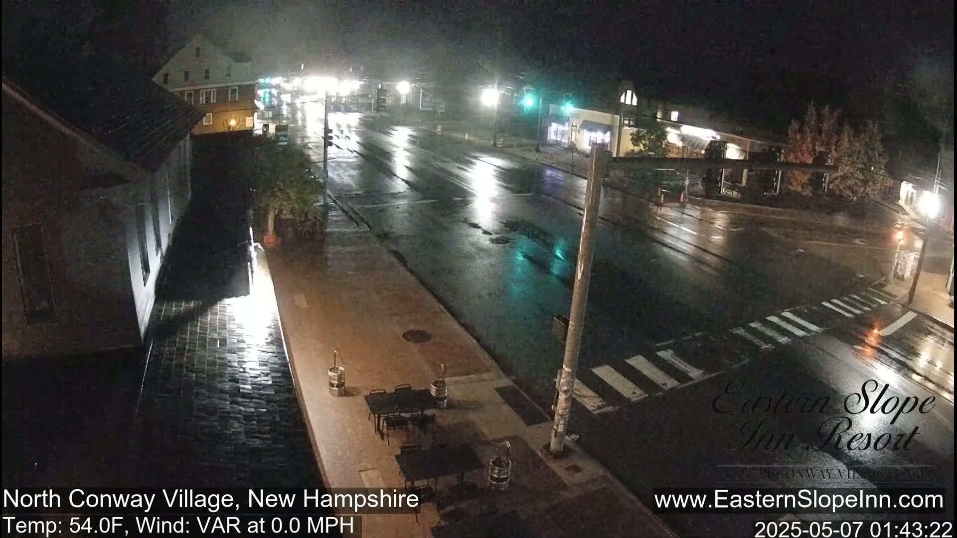 A rain-slicked street scene at night shows buildings and empty outdoor seating under streetlights.