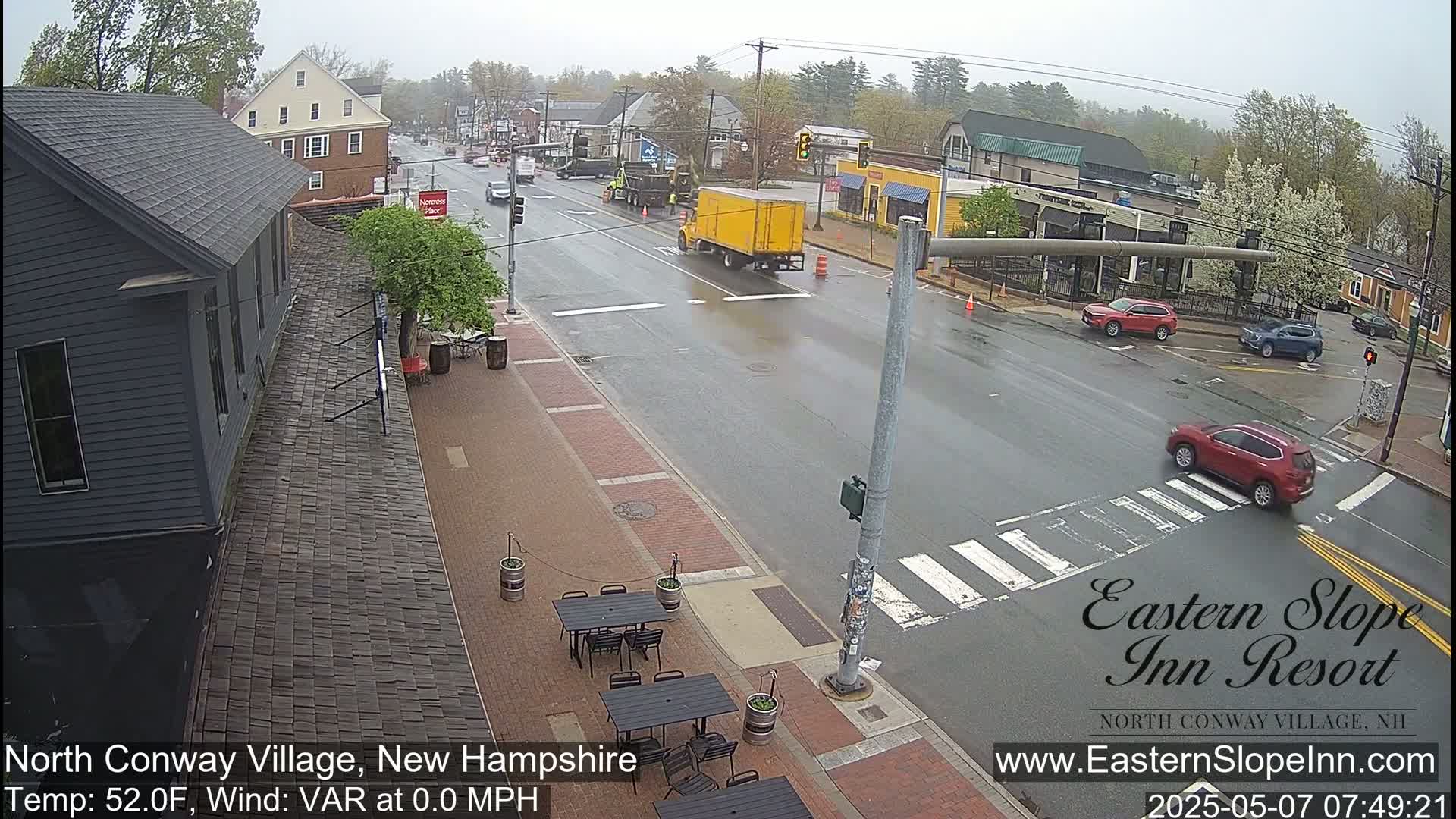 A rainy street scene shows several cars at an intersection, a yellow delivery truck, and outdoor seating in front of a dark gray building.