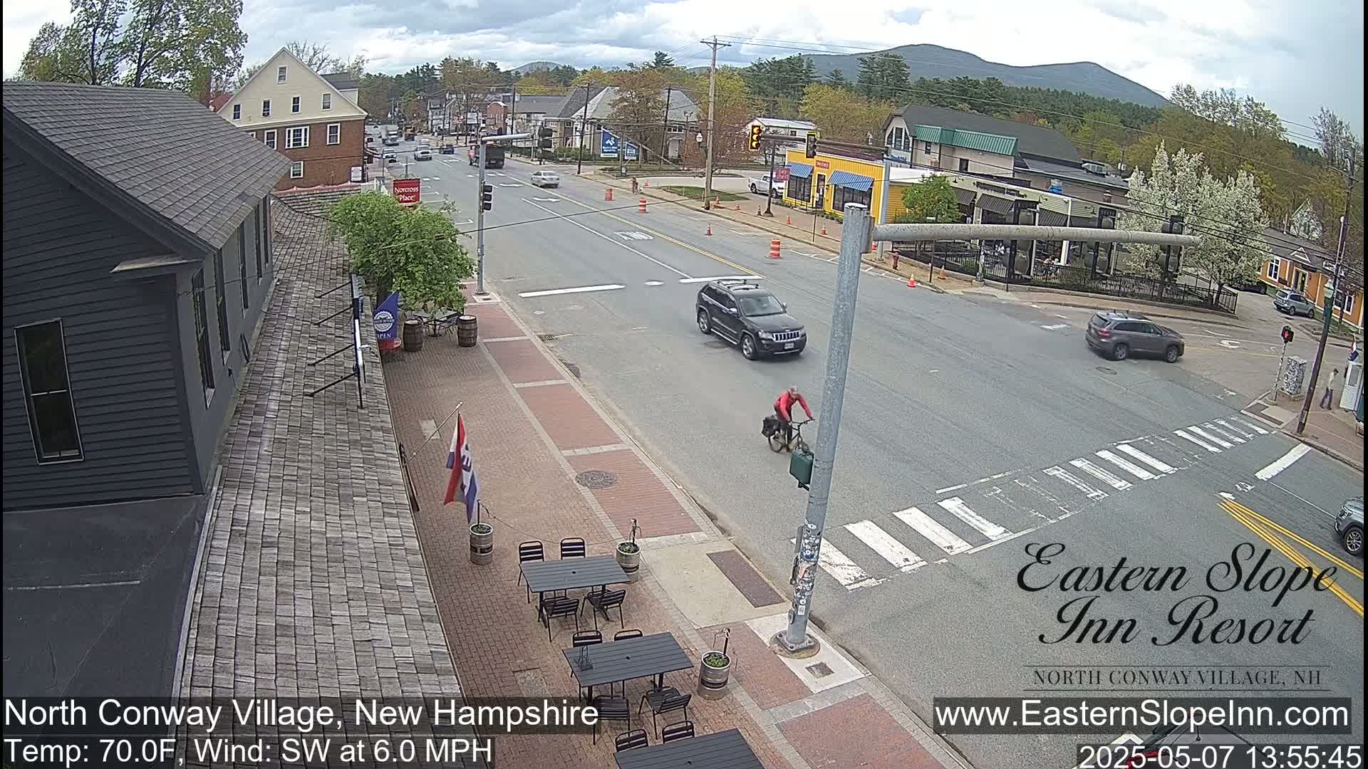 A cyclist rides past a street intersection in a village, with cars driving by and outdoor seating at a restaurant, under partly cloudy skies.