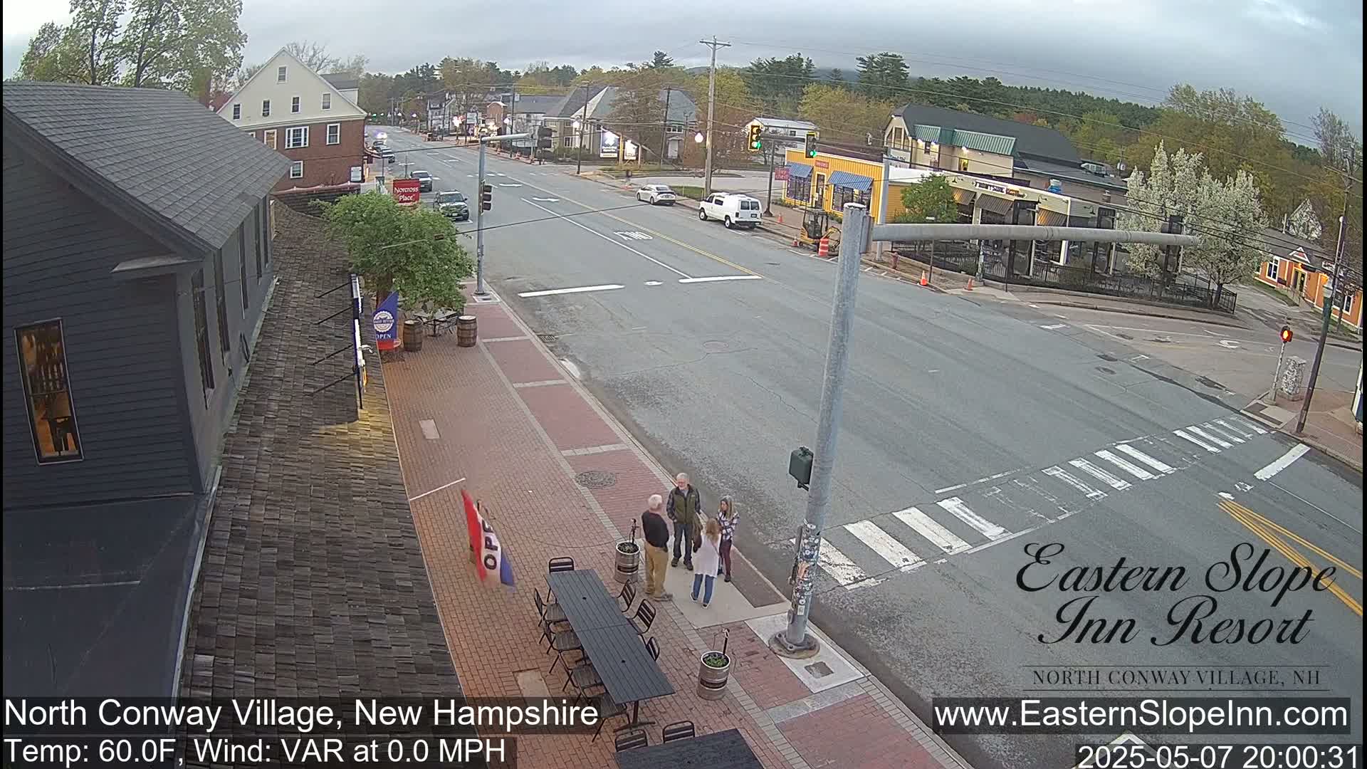 A street scene in a small town on an overcast day shows a few pedestrians, several cars, and buildings including a dark-colored building with a visible awning, a yellow building, and others lining the street.