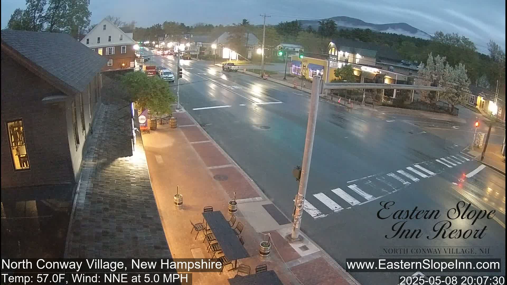 A wet, nighttime street scene shows a village with low-rise buildings and some cars under a partly cloudy sky.