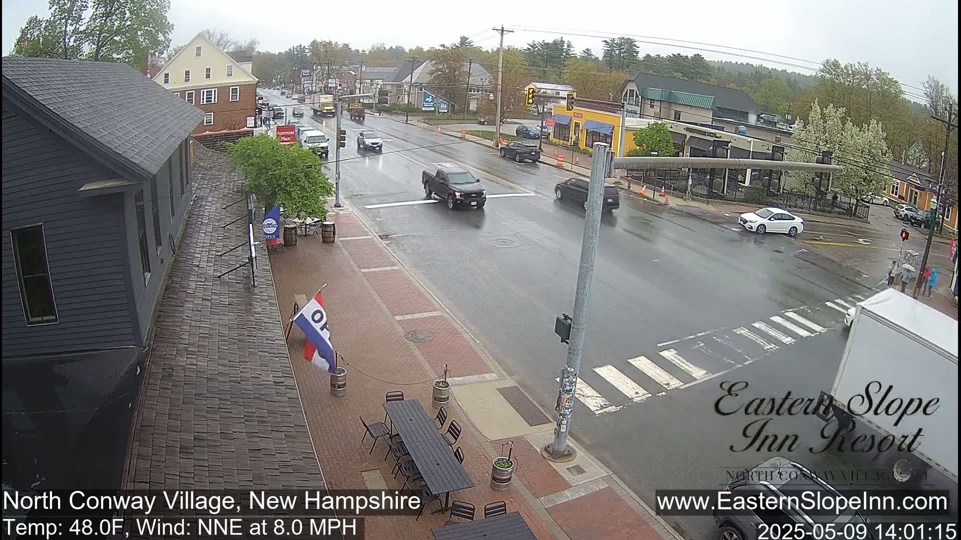 A rain-slicked street scene shows several vehicles at an intersection in front of buildings and outdoor seating areas.