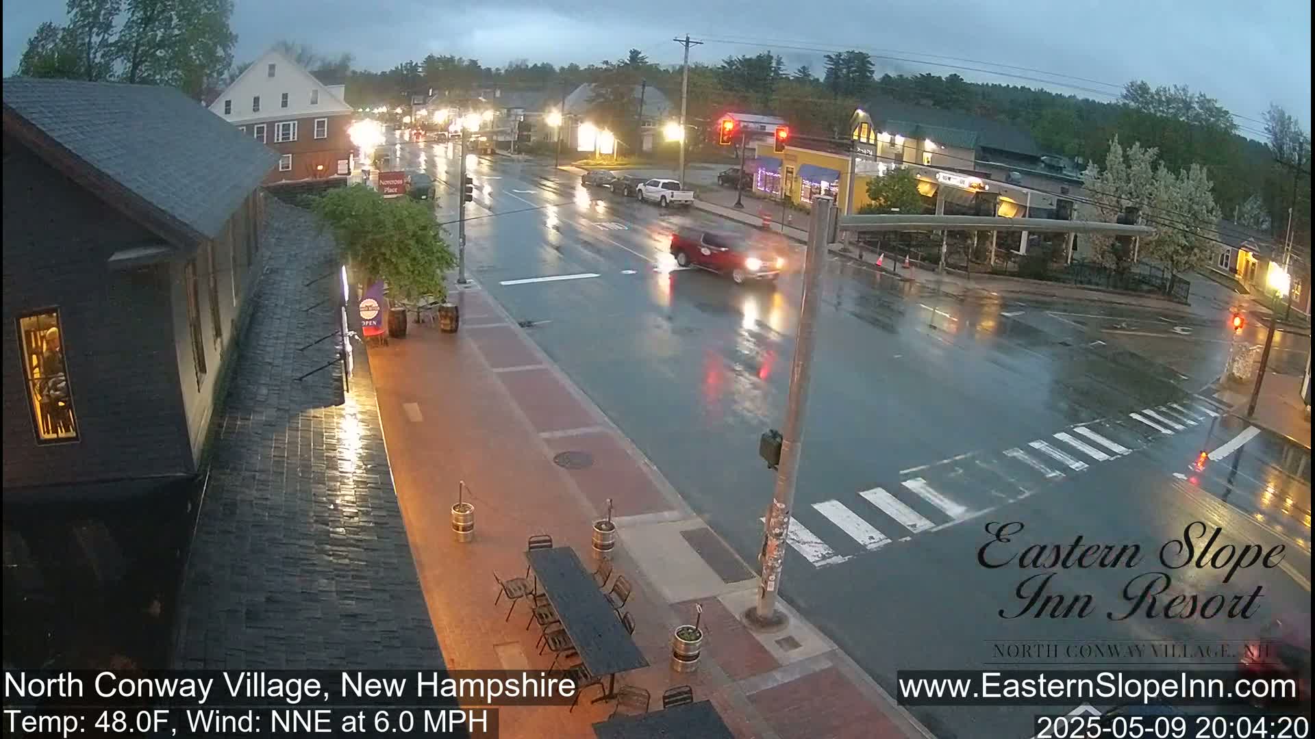 A rain-slicked street scene at night shows a few cars driving through a town intersection, with outdoor seating at a restaurant visible to the side.