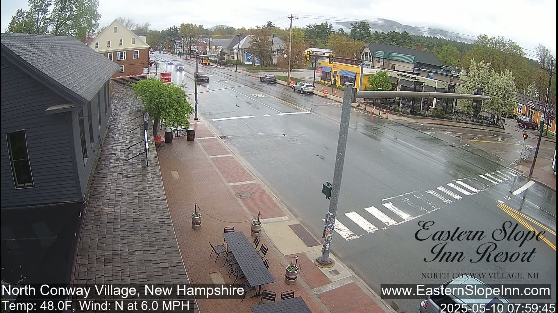 A wet street scene in a village shows several cars driving on a road with a crosswalk, and an outdoor patio with tables and chairs sits near a dark-grey building.