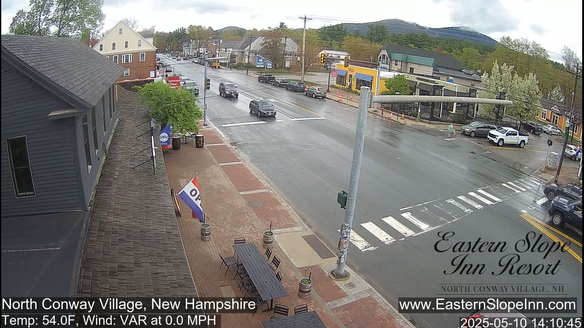 A wet street scene shows several cars driving through a town with buildings lining the road, under a cloudy sky.