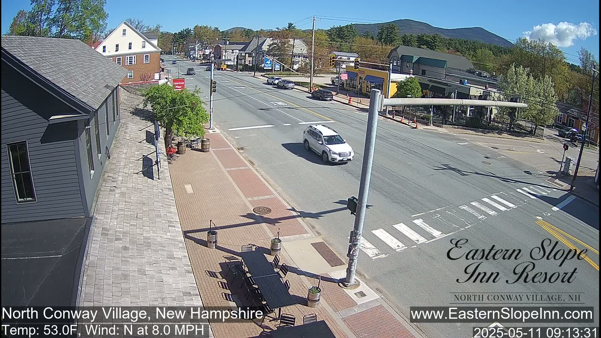 A sunny street scene shows a paved road with several cars and a pedestrian crosswalk, alongside buildings and outdoor seating in a village setting.