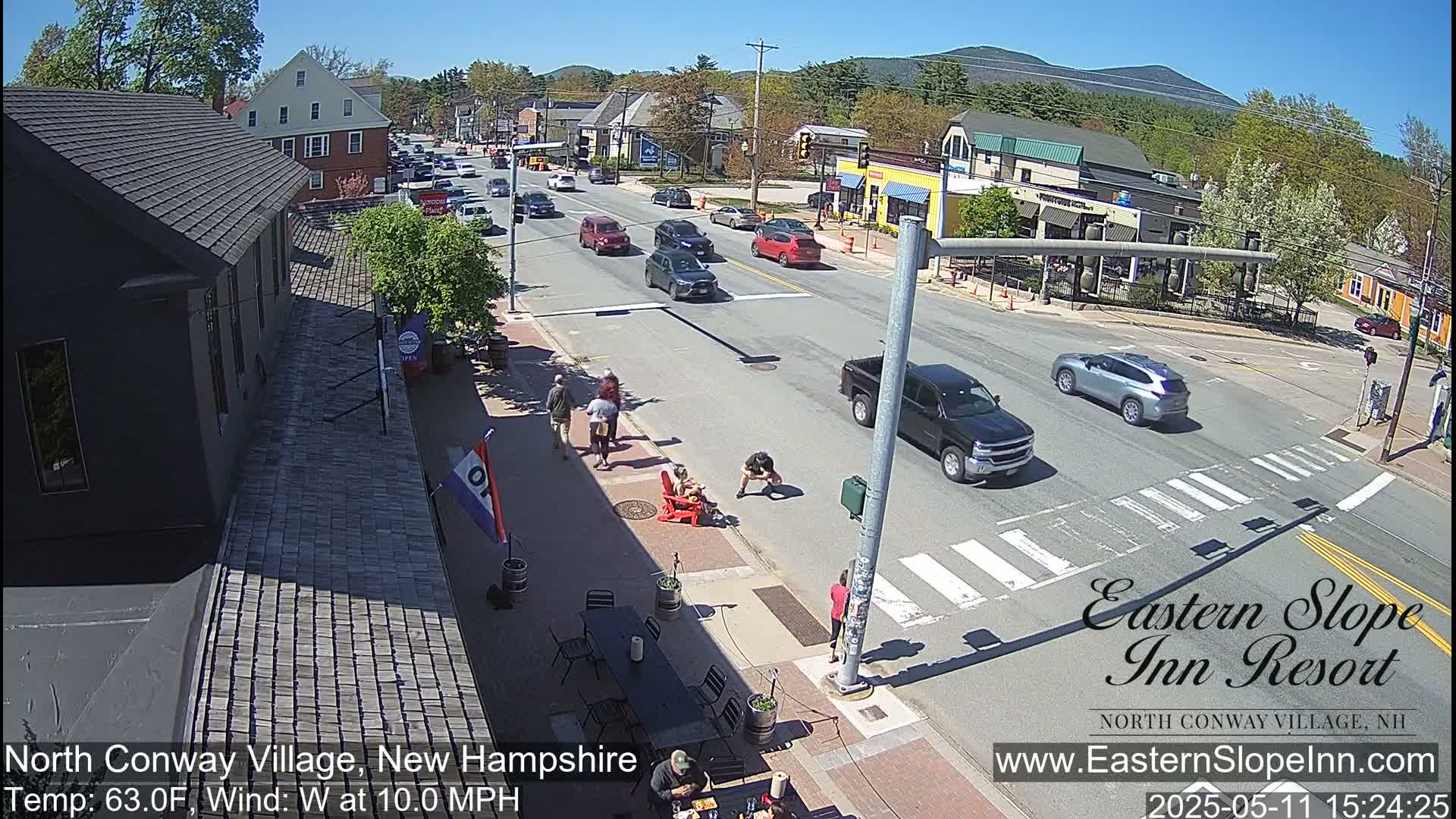 A sunny street scene shows several cars driving on a road with pedestrians walking on the sidewalk, and some people sitting at outdoor tables.
