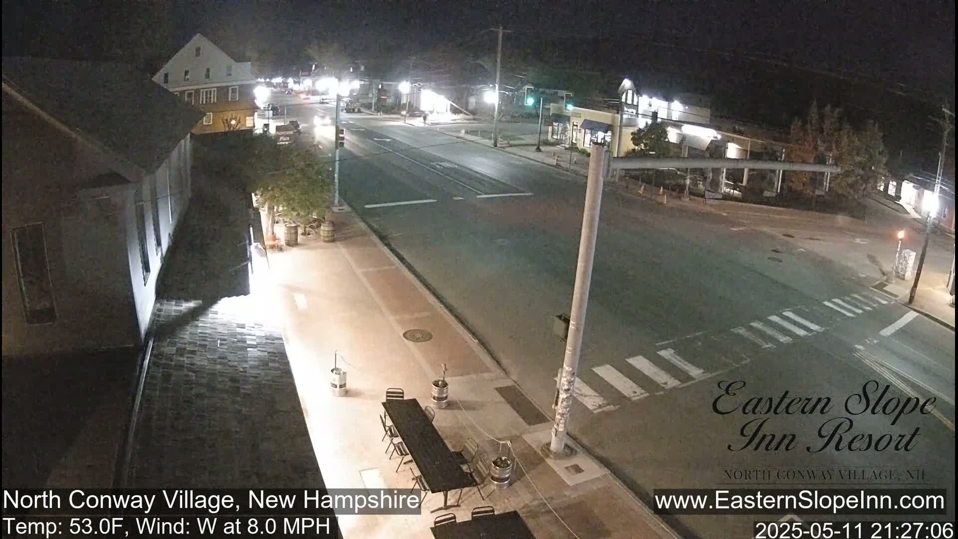 A nighttime view of a mostly empty street intersection in a small town, with a few cars visible and a calm, cool breeze.
